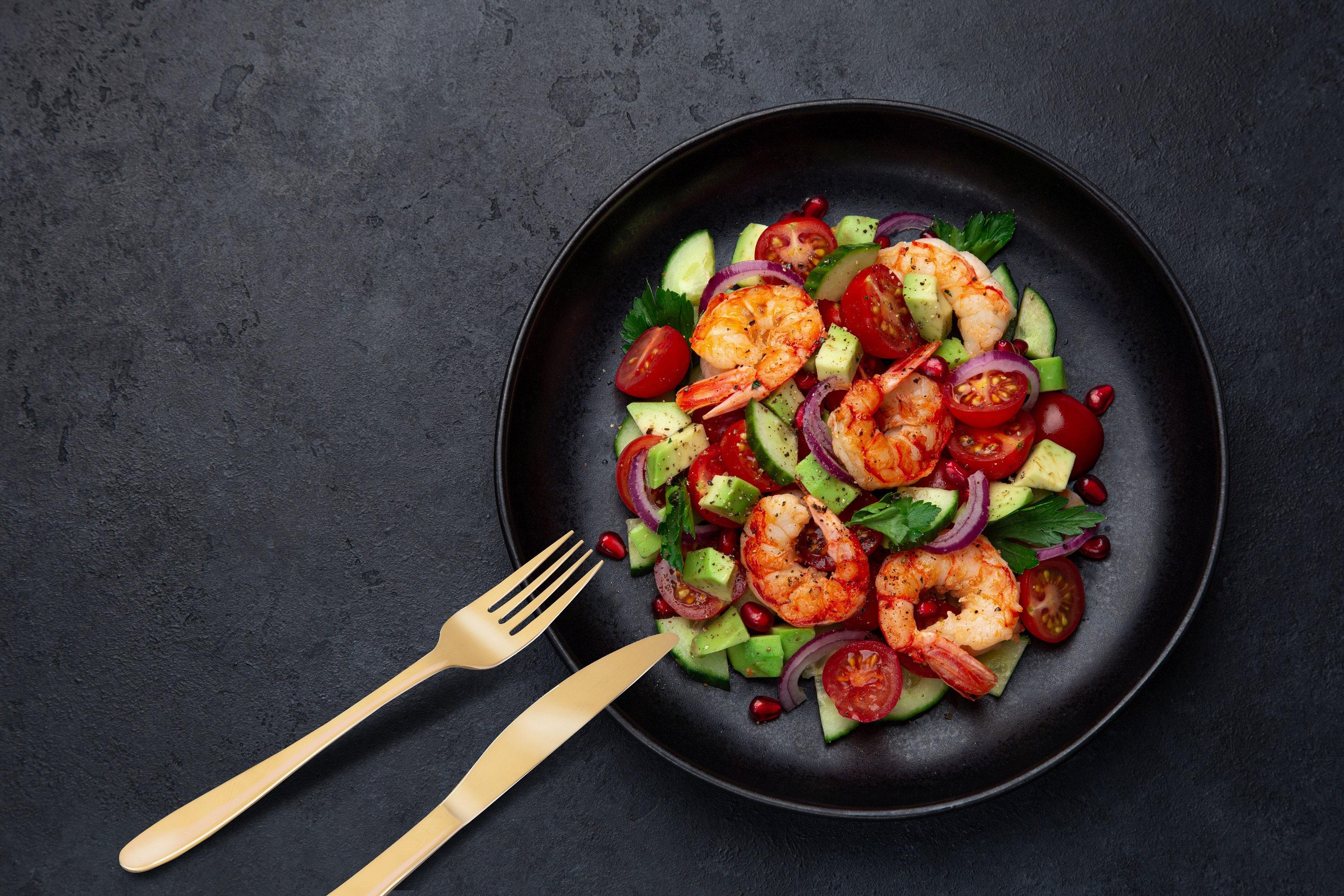 Shrimp salad with tomatoes, avocado, and red onion on a black plate, with golden fork and knife.