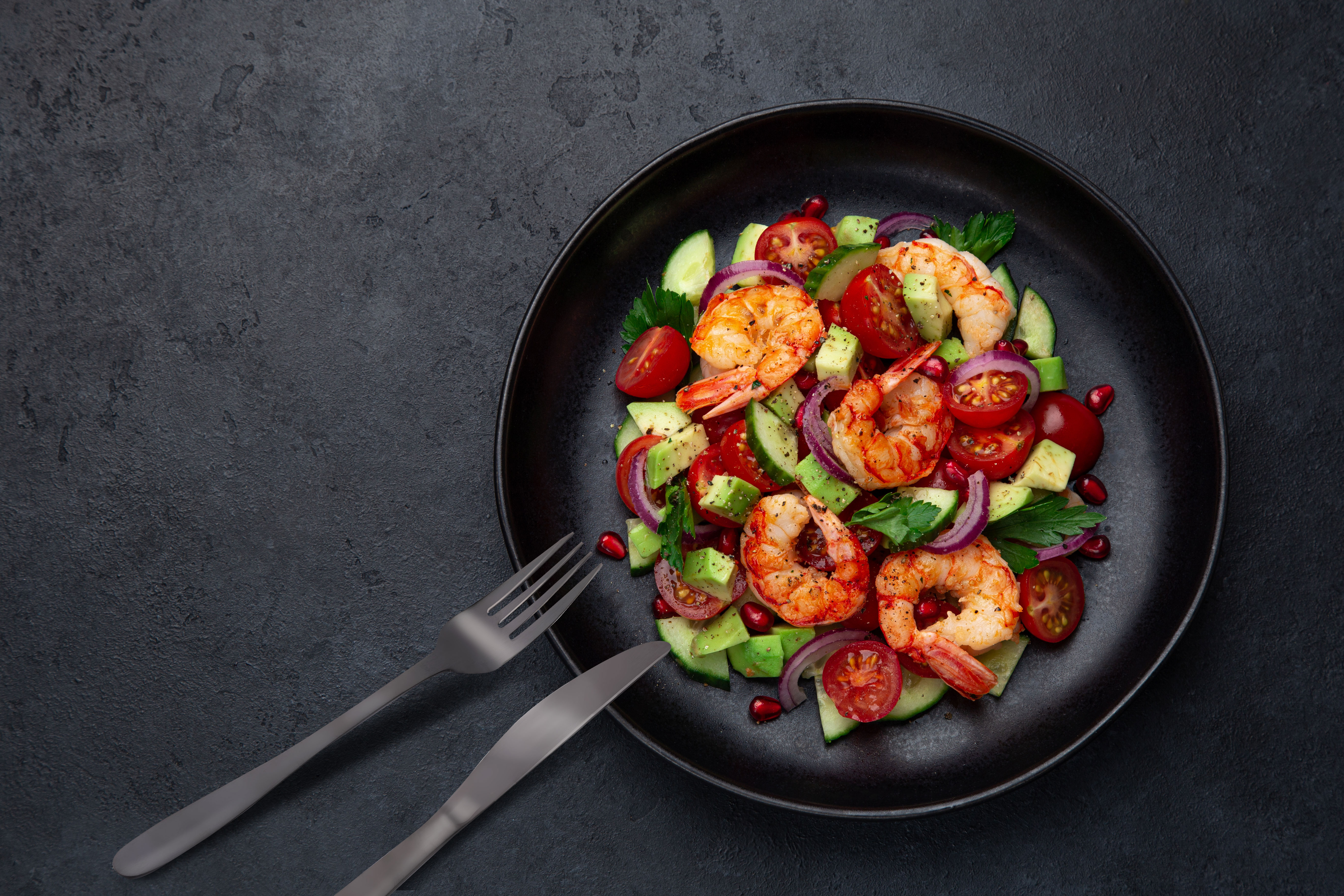 A shrimp salad with tomatoes, cucumber, red onion, and avocado on a black plate. Fork and knife on the side.