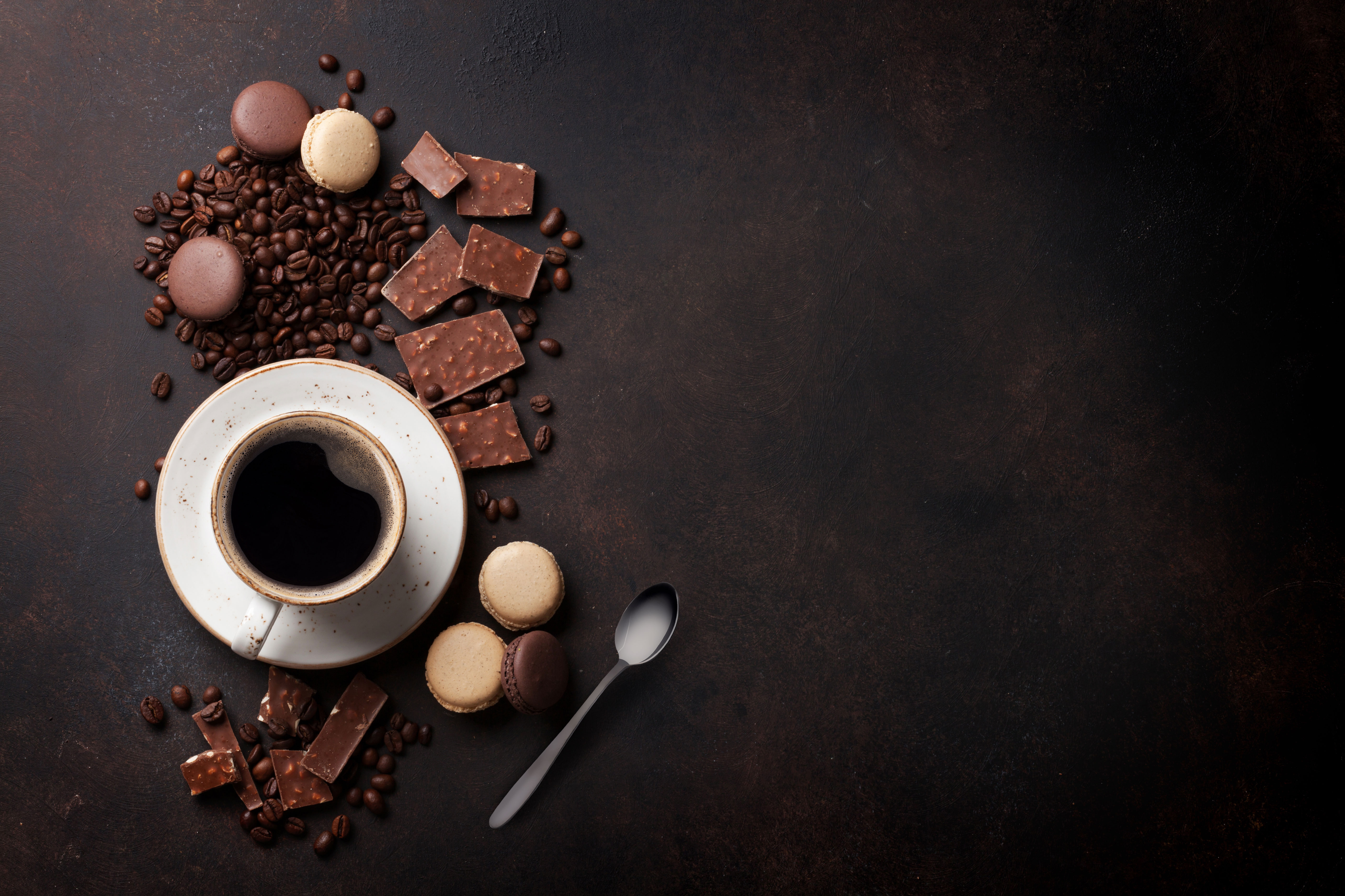 Overhead shot of coffee, chocolate, macaroons, and coffee beans on a dark surface. A spoon is also visible.