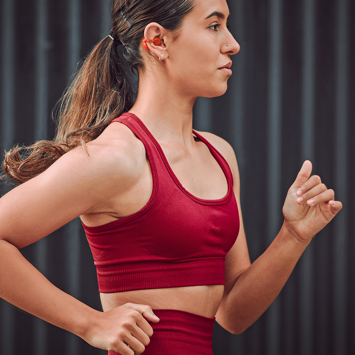 Kobieta biegnie w czerwonym stroju sportowym i słuchawkach. Jej włosy są związane w kucyk. Tło to ciemna, teksturowana ściana.