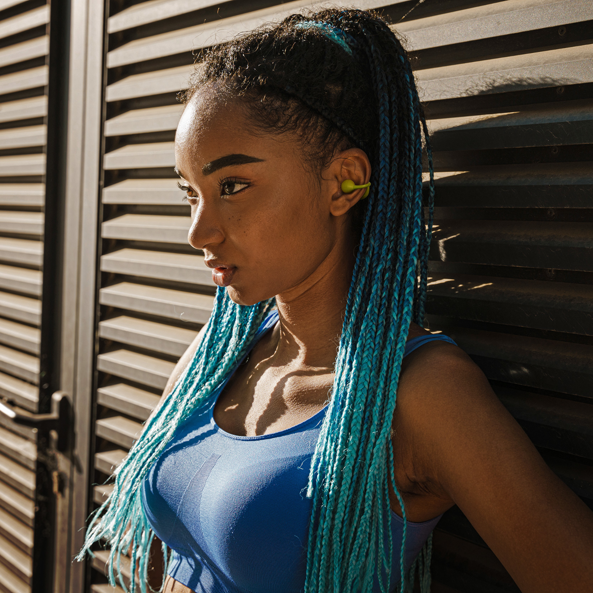 Kobieta z niebieskimi warkoczami w niebieskim topie i żółtych słuchawkach, opierająca się o ciemną ścianę z listew.