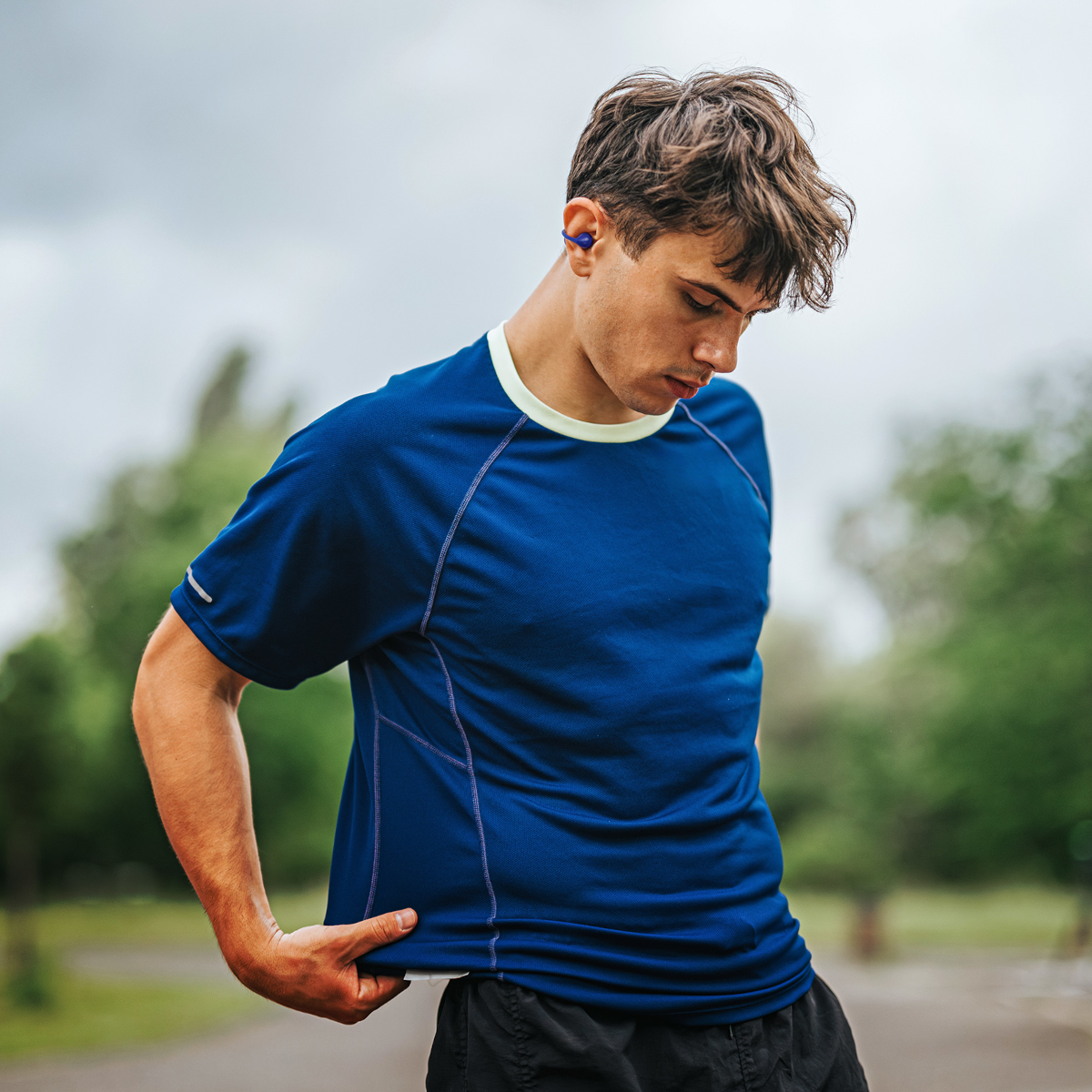 Mężczyzna w niebieskiej koszulce i czarnych szortach patrzy w dół. Ma słuchawki i stoi na ścieżce z drzewami w tle.