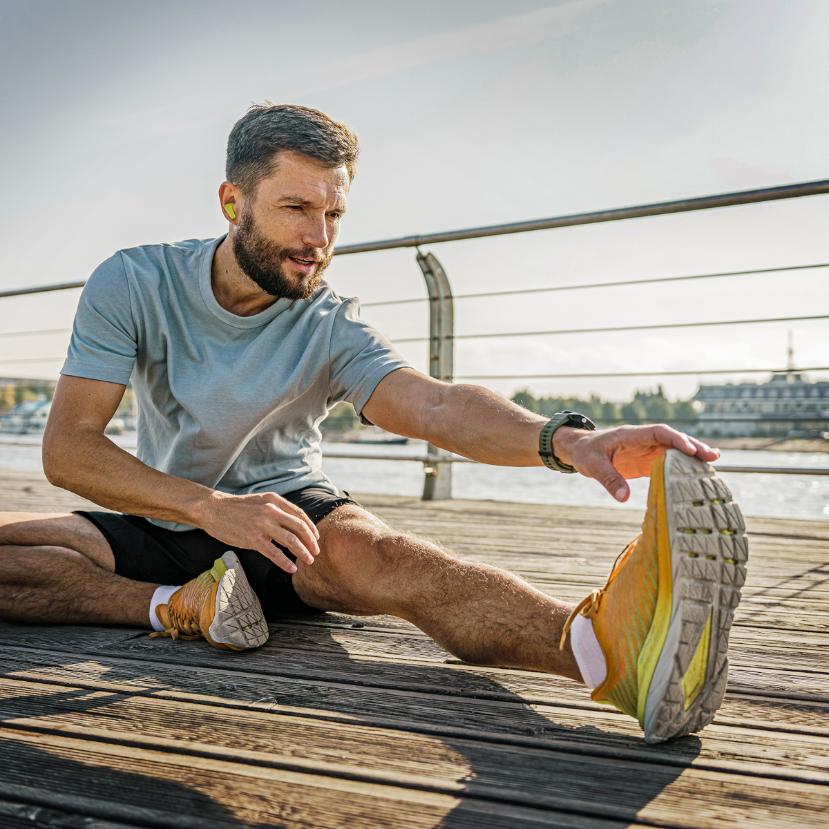 Mężczyzna rozciąga nogę na drewnianym molo. Ma na sobie szarą koszulkę i czarne szorty. W tle budynki i woda.