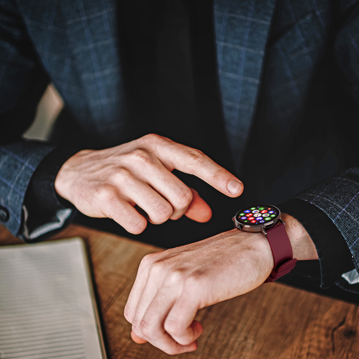Un uomo in giacca e cravatta indica uno smartwatch con cinturino bordeaux. Il quadrante mostra icone di app colorate.