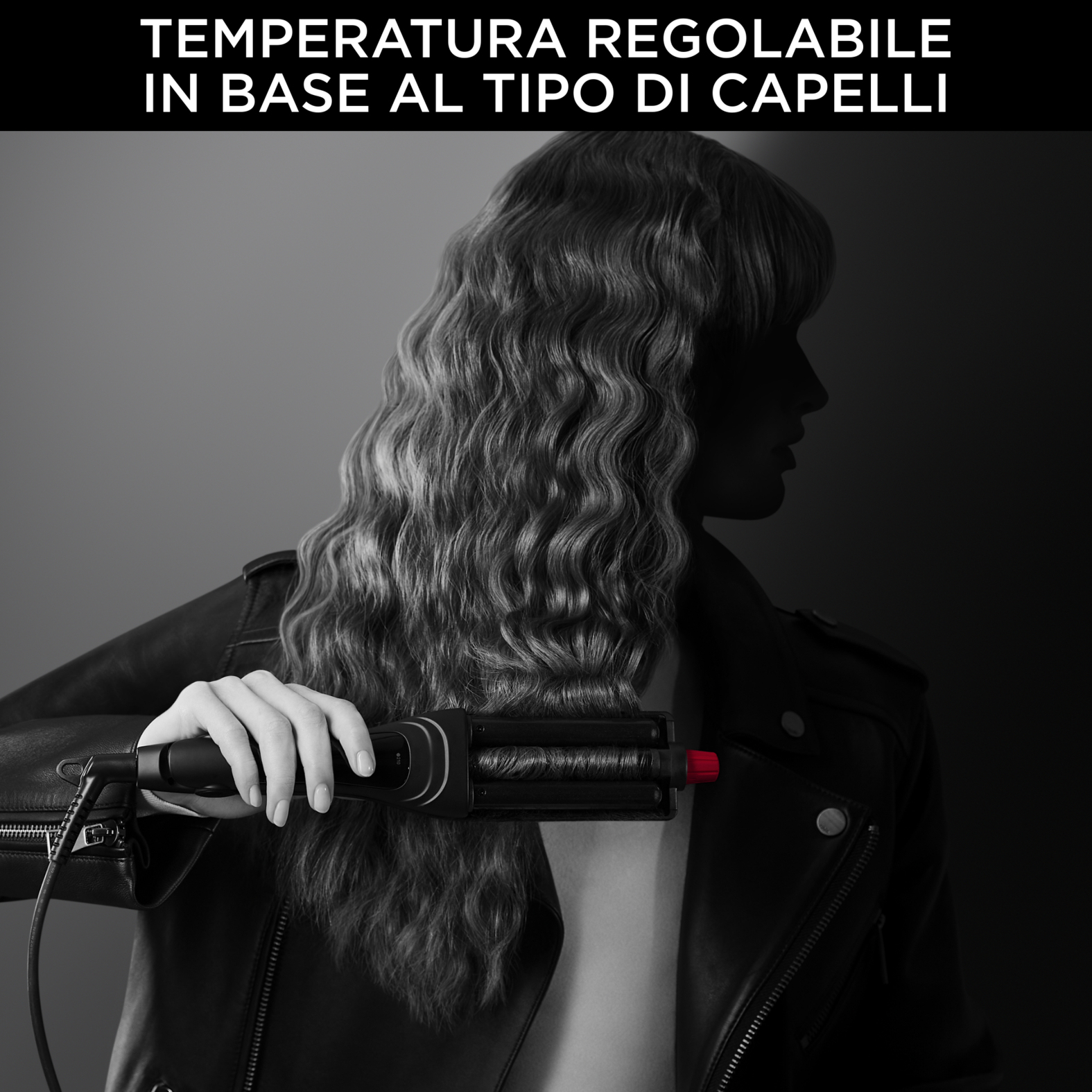 Eine Frau hält ein schwarzes Haarstyling-Werkzeug. Sie hat welliges Haar und trägt eine schwarze Lederjacke. Der Hintergrund ist dunkel.