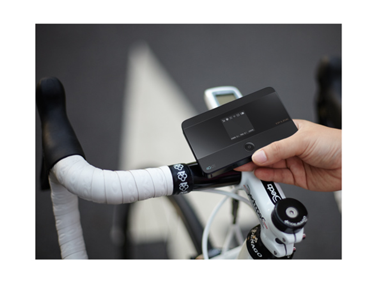 Eine Hand hält ein schwarzes Gerät über einem Fahrradlenker. Der Lenker hat weißes Band und einen schwarzen Griff. Ein Tacho ist sichtbar.