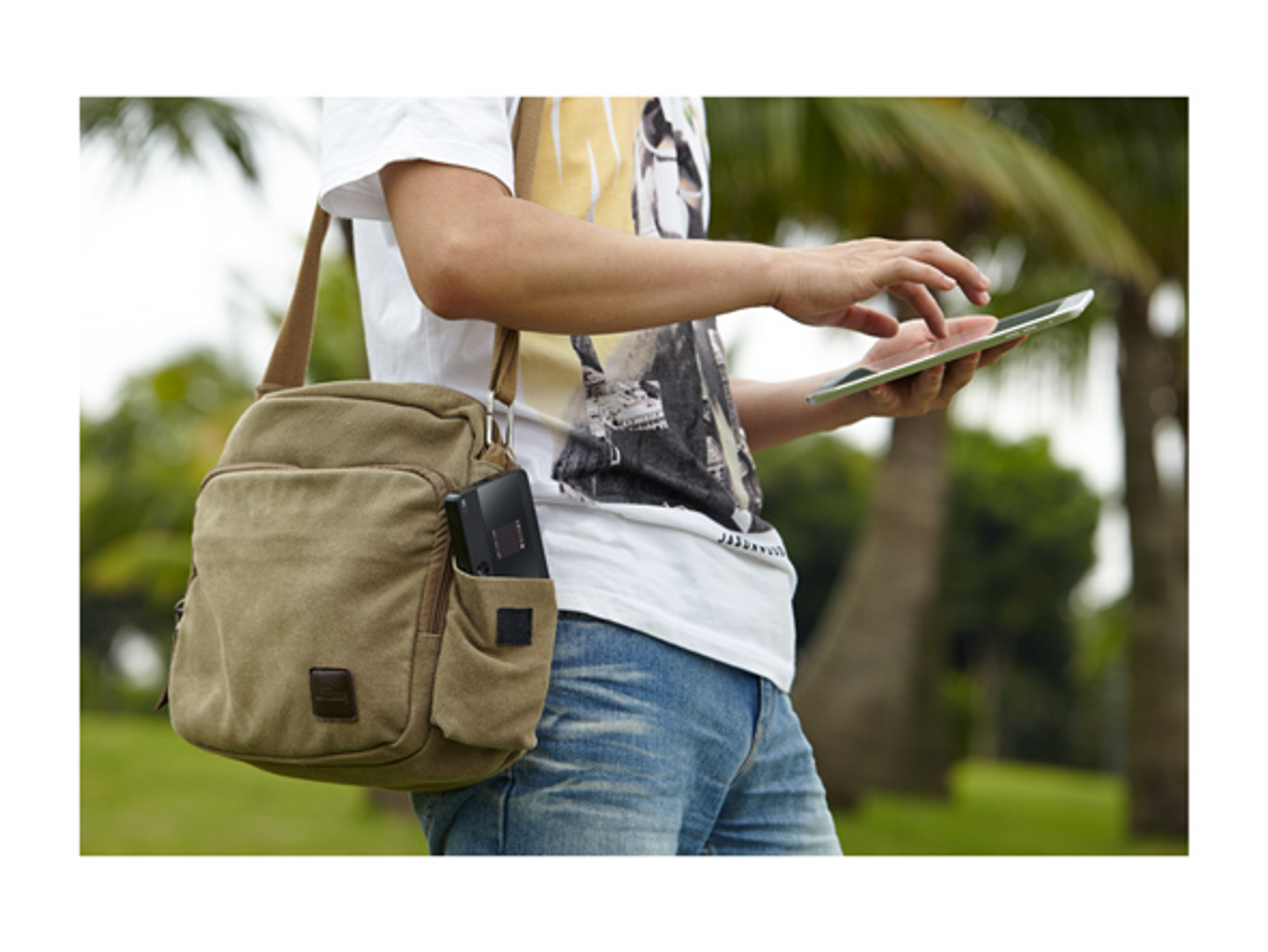 Eine Person benutzt ein Tablet. Sie trägt ein weißes Hemd, Jeans und trägt eine Tasche. Palmen sind im Hintergrund.