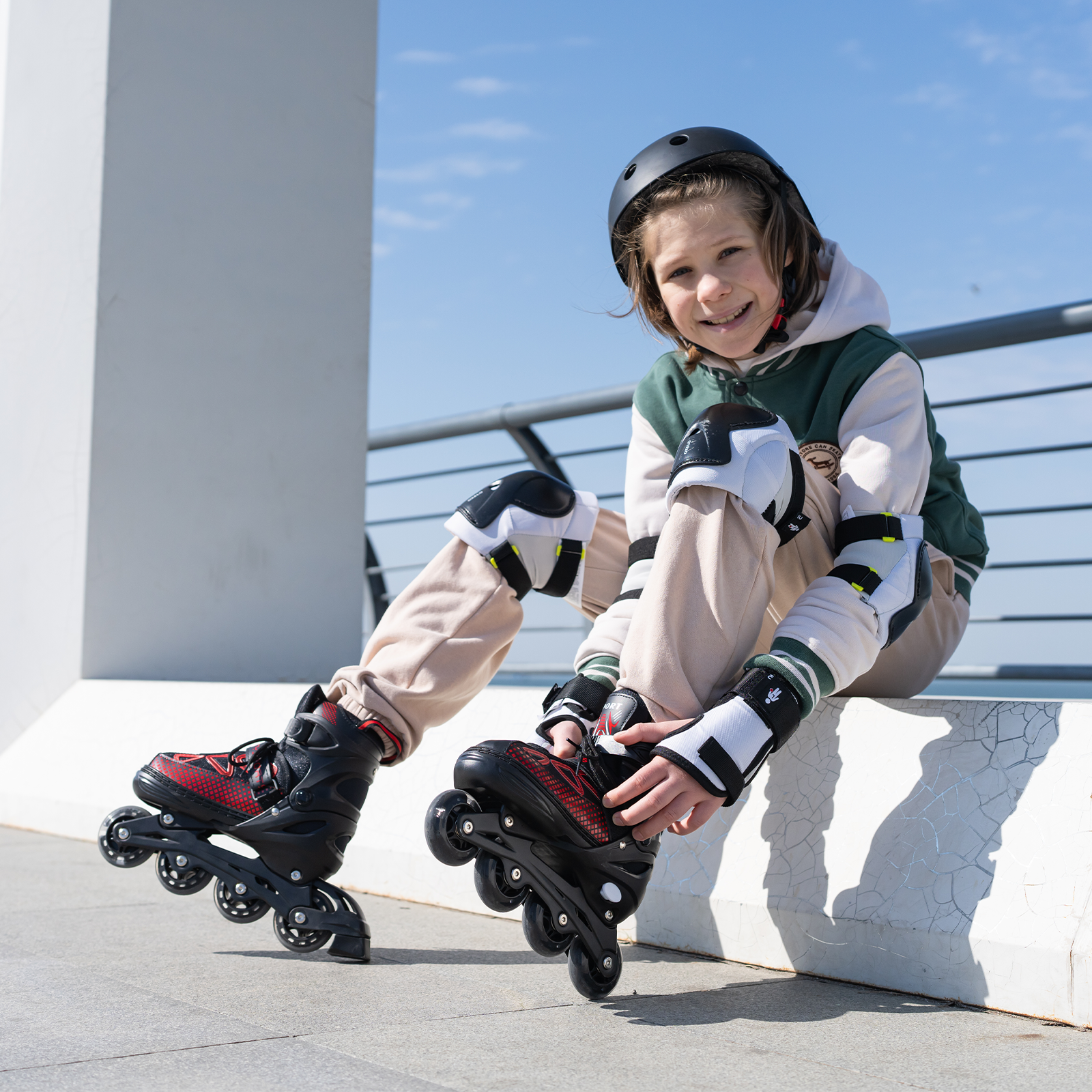 Un niño con casco y equipo de protección se sienta, poniéndose patines. El cielo es azul.