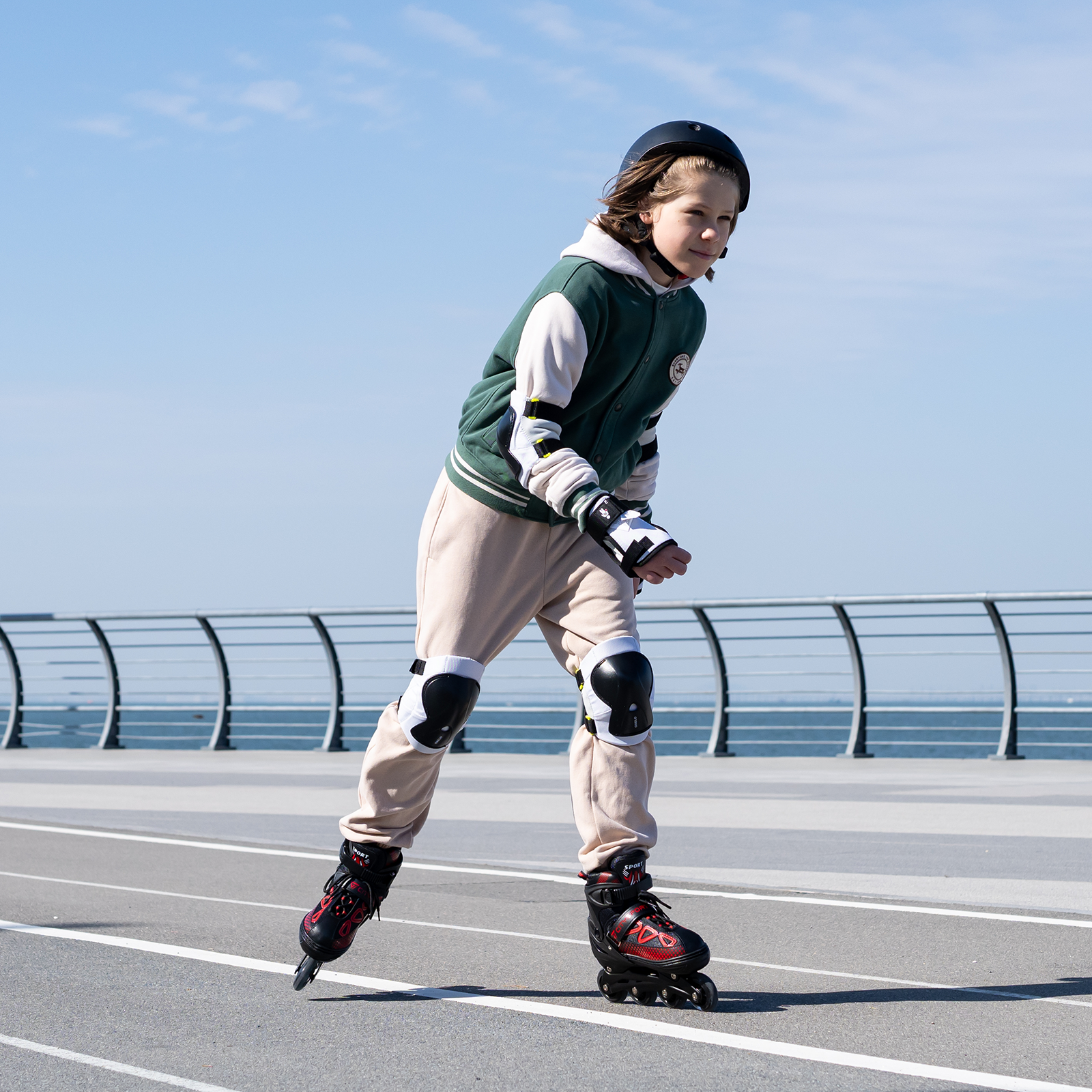 Una persona en patines, con casco y protecciones. Están en una pista con barandilla y el mar de fondo.