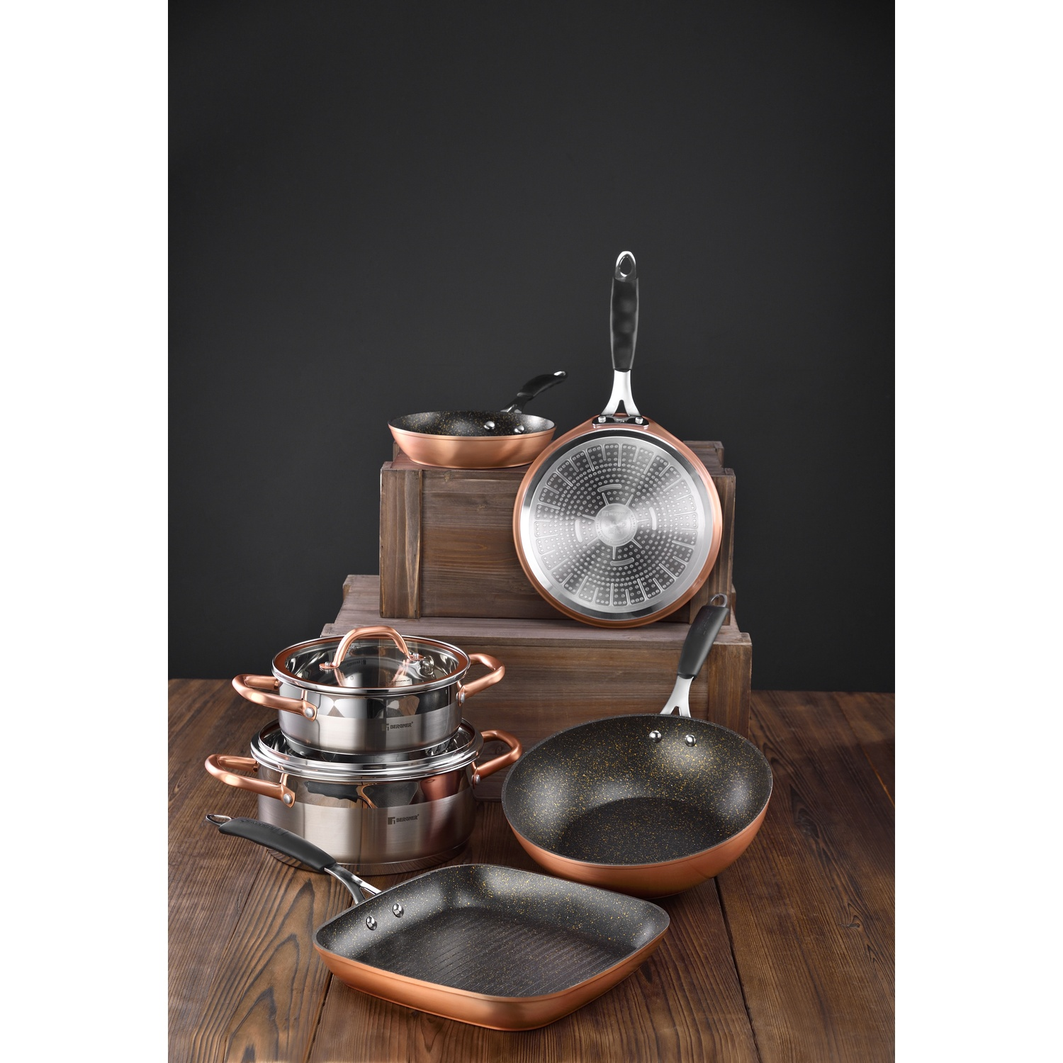 Juego de utensilios de cocina sobre una superficie de madera. Incluye ollas, sartenes y tapas con detalles en cobre y negro. Fondo oscuro y cajas de madera.