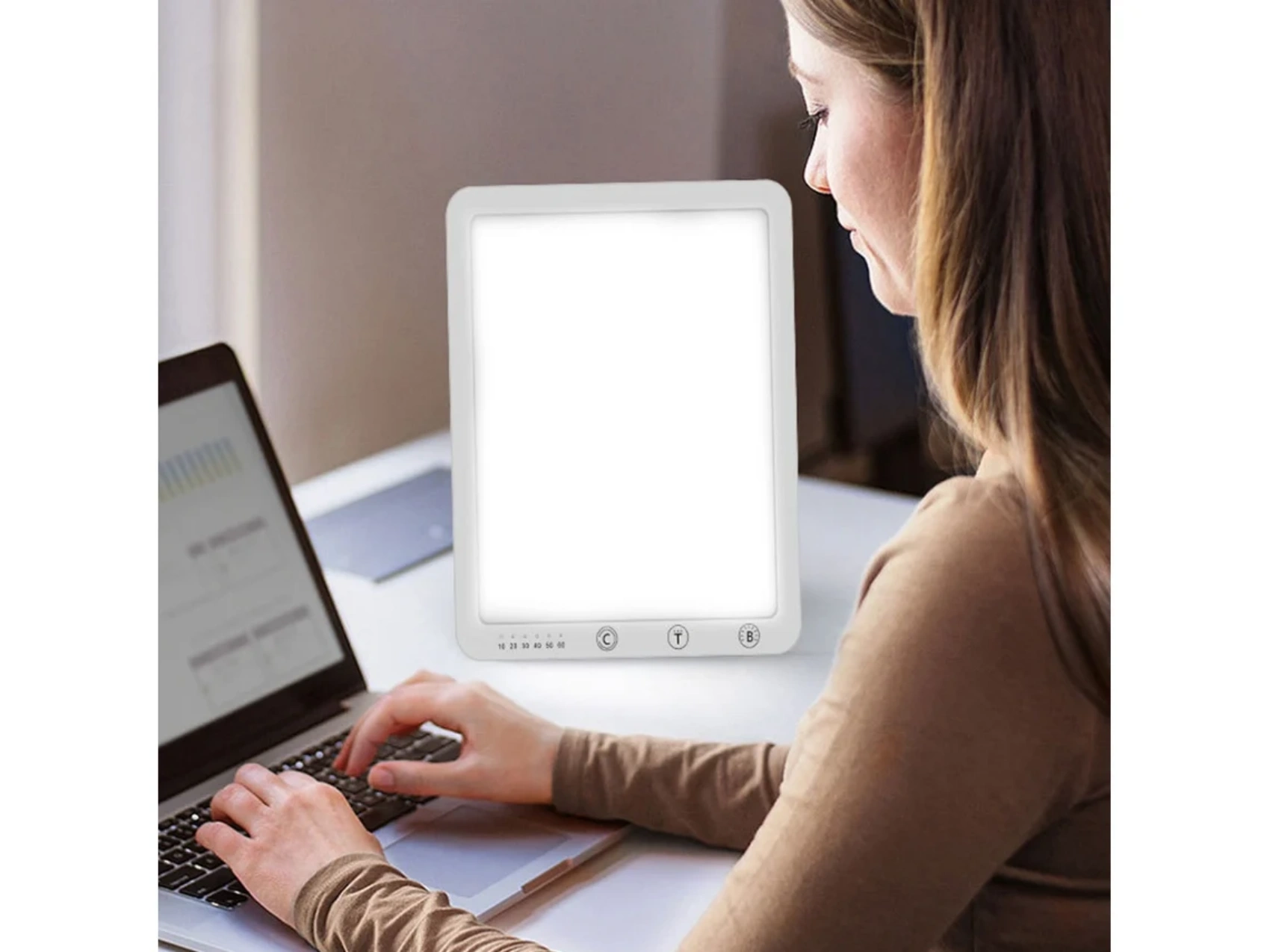 Eine Frau benutzt einen Laptop mit einer hellen Lichttherapielampe. Die Lampe steht auf einem weißen Tisch. Sie trägt ein braunes Hemd.