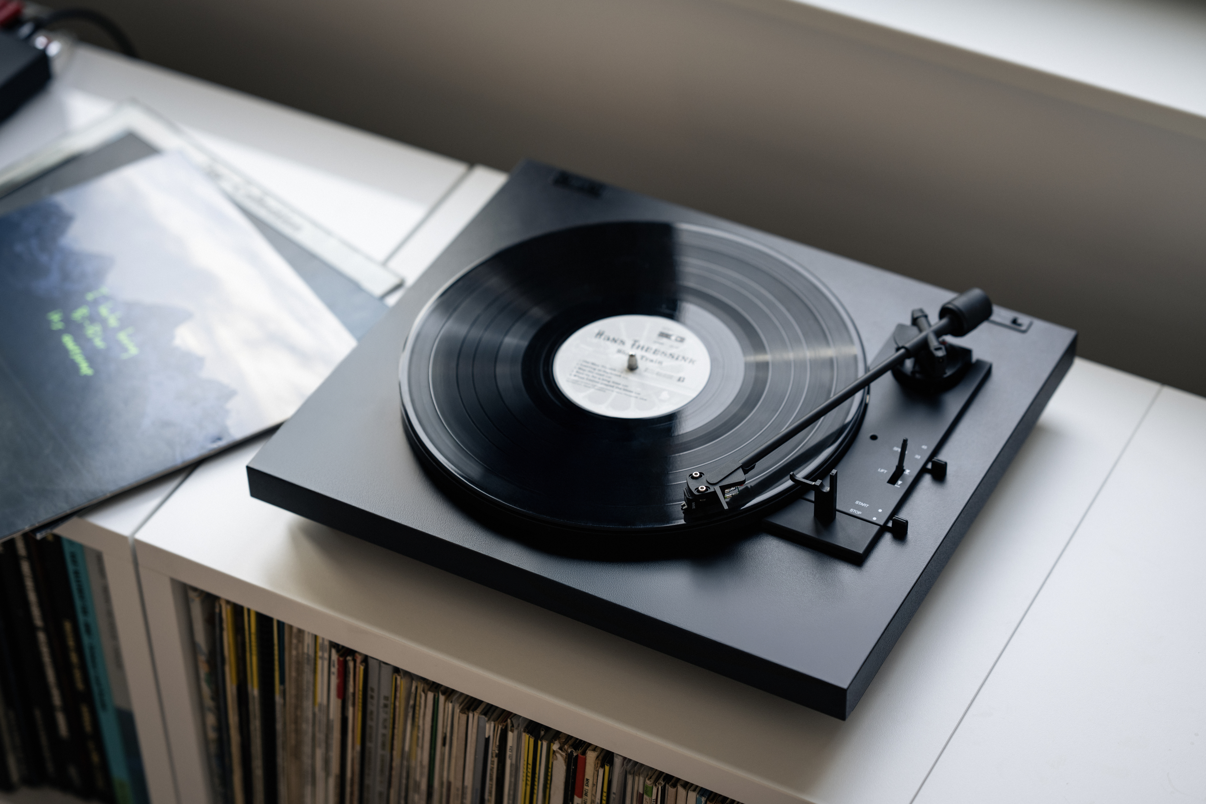 Een platenspeler met een zwarte plaat die draait. De platenspeler staat op een witte plank met albums. Een tweede album ligt ernaast.