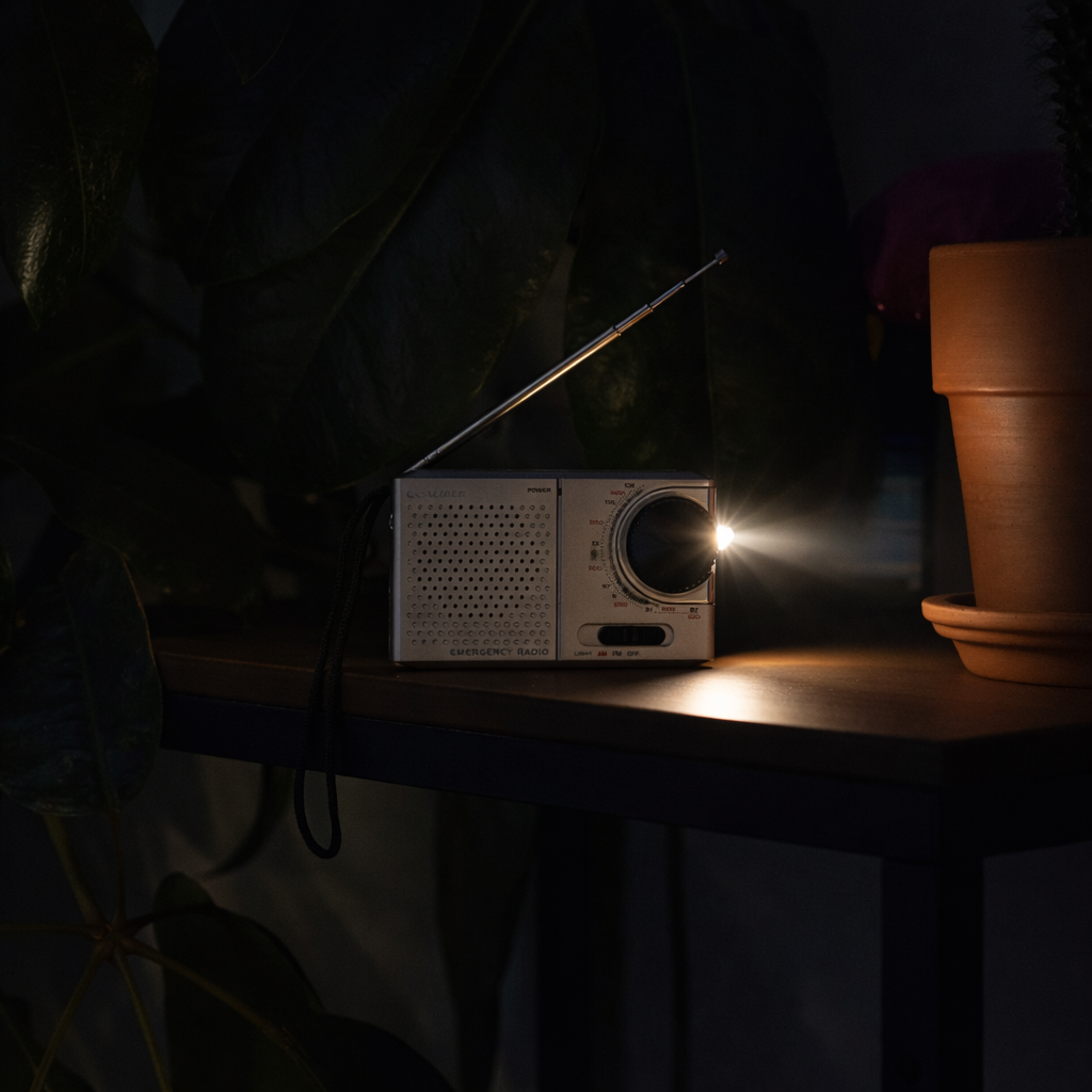 Zilveren noodradio met antenne verlicht door een helder licht. Naast een terracotta pot, tegen een donkere groene achtergrond.