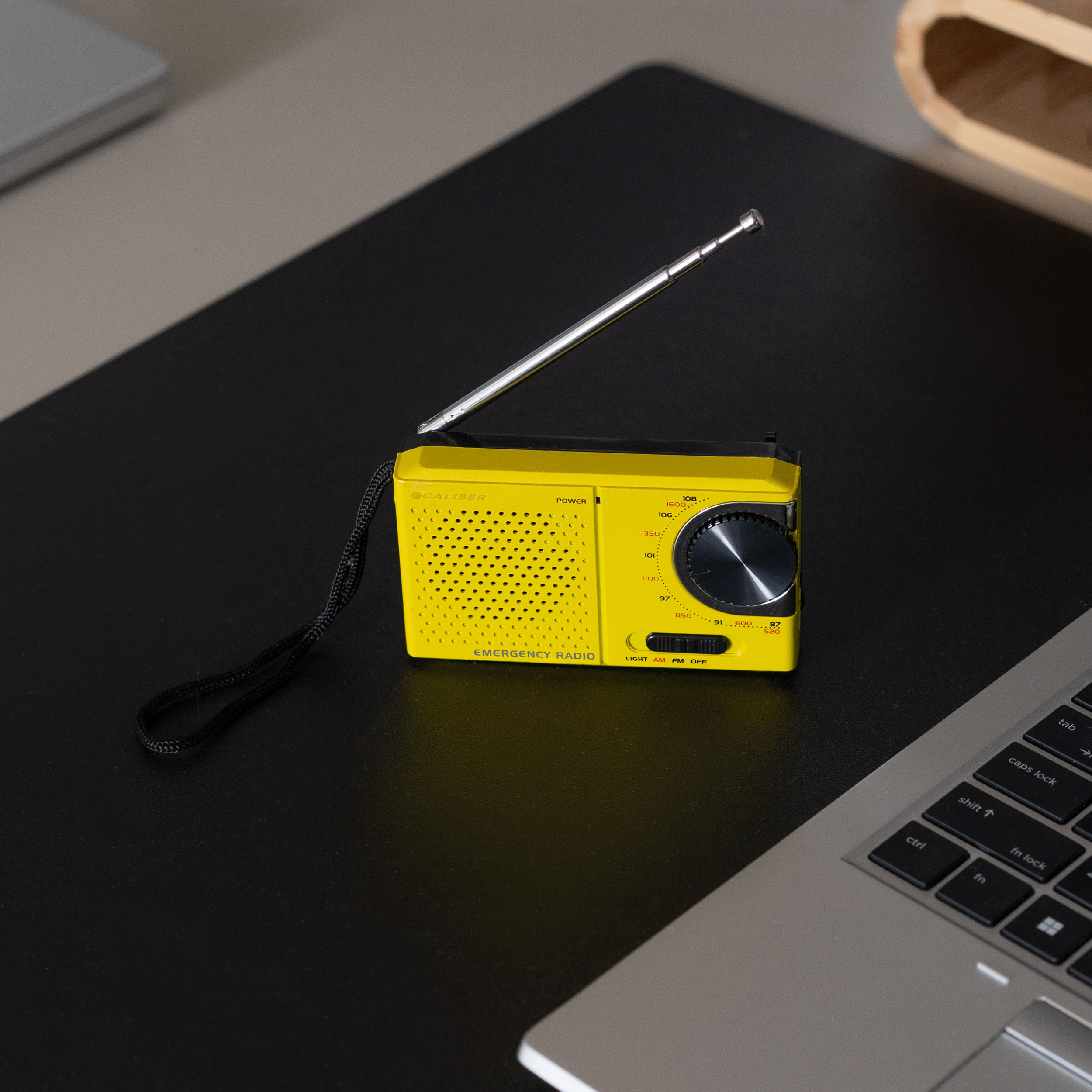 A yellow emergency radio with an antenna sits on a black surface next to a laptop.