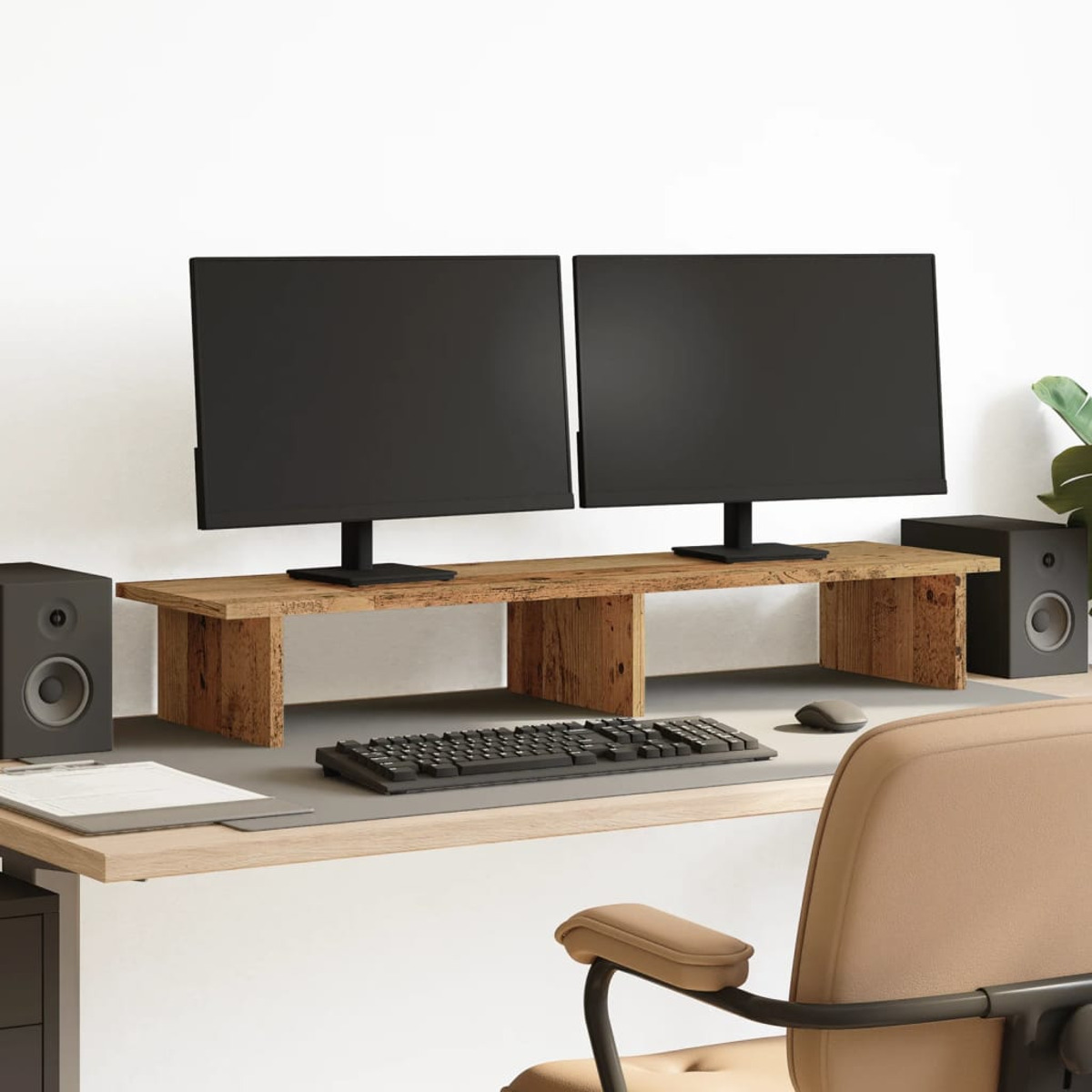 Ein Schreibtisch mit zwei Monitoren auf einem Holzständer, Lautsprechern, Tastatur, Maus und einem Stuhl. Weiße Wand im Hintergrund.