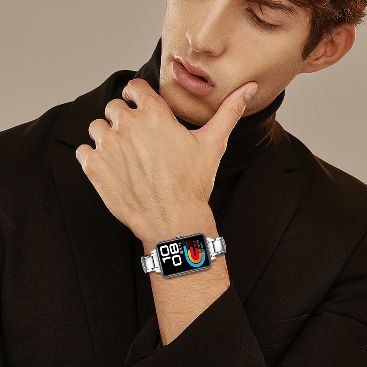 Ein Mann im schwarzen Anzug und Rollkragen, der sein Gesicht berührt. Eine Smartwatch mit farbenfrohem Display an seinem Handgelenk.