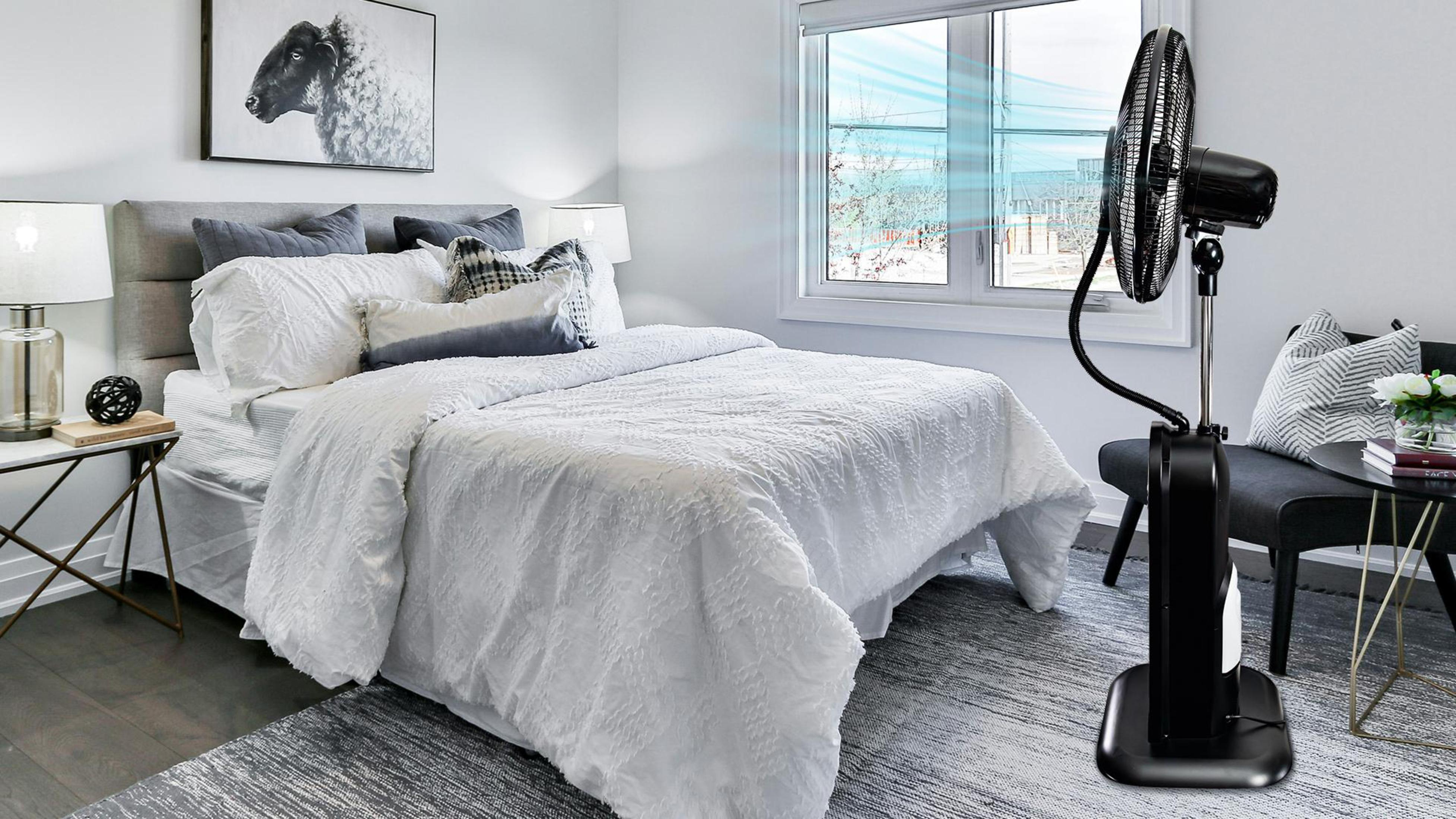 Schlafzimmer mit Bett, Ventilator und Fenster. Ein schwarzer Ventilator bläst Luft. Ein Bild eines Schafs hängt an der Wand.