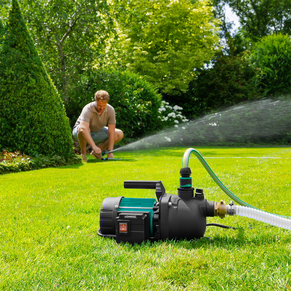 Een man hurkt en stelt een sproeier af. Een waterpomp staat op de voorgrond. Groen gras en bomen vullen de achtergrond.