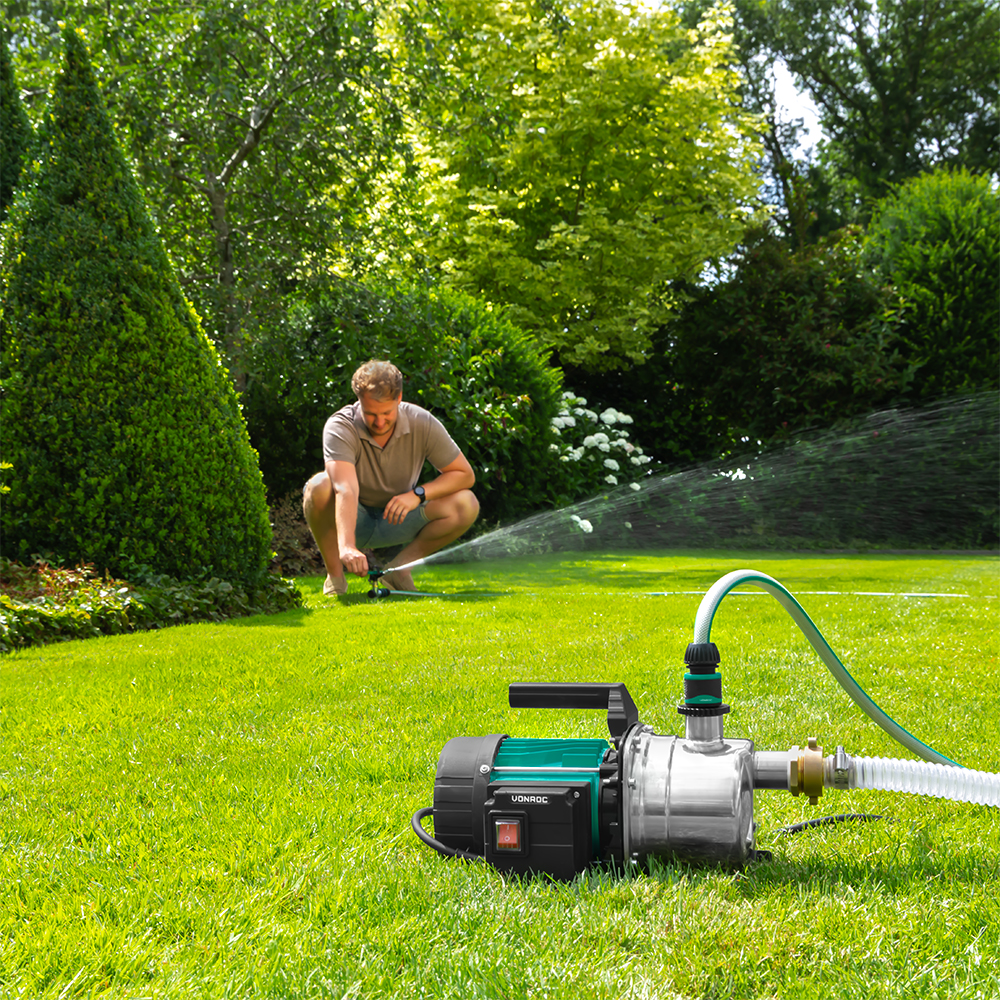Een man past een sproeier aan in een groene tuin. Een waterpomp staat op de voorgrond. Bomen en struiken vullen de achtergrond.