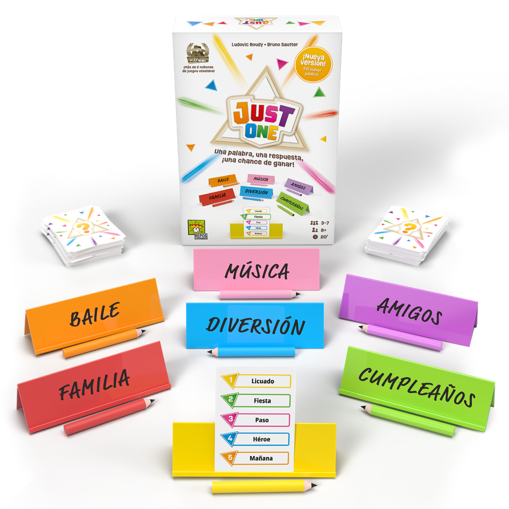 Se muestra el juego de mesa 'Just One'. Incluye cartas, letreros de colores con palabras, lápices y la caja del juego en el fondo.