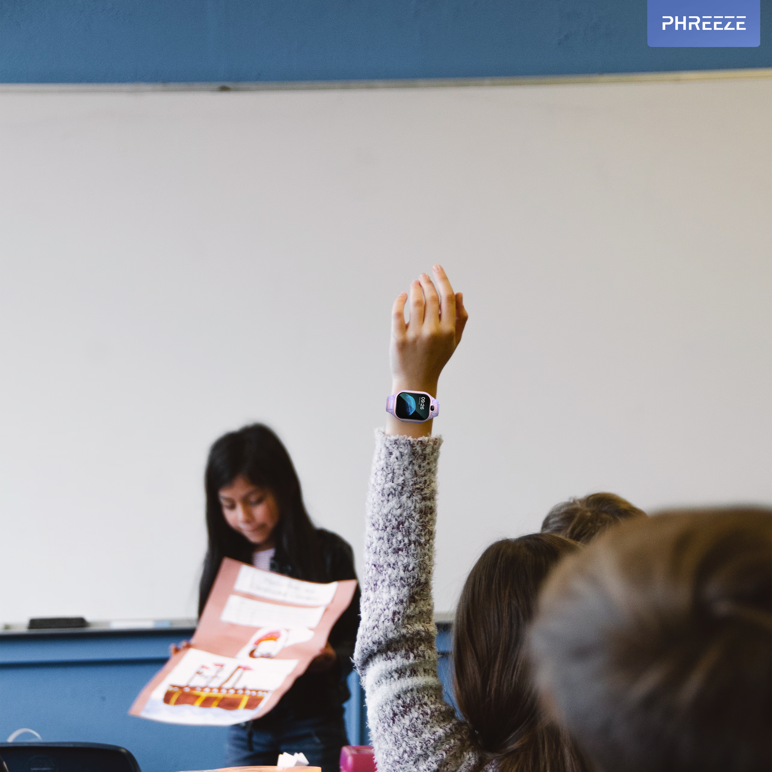 Een leerling steekt een hand op met een smartwatch. Een andere leerling presenteert. Klaslokaal met whiteboard.