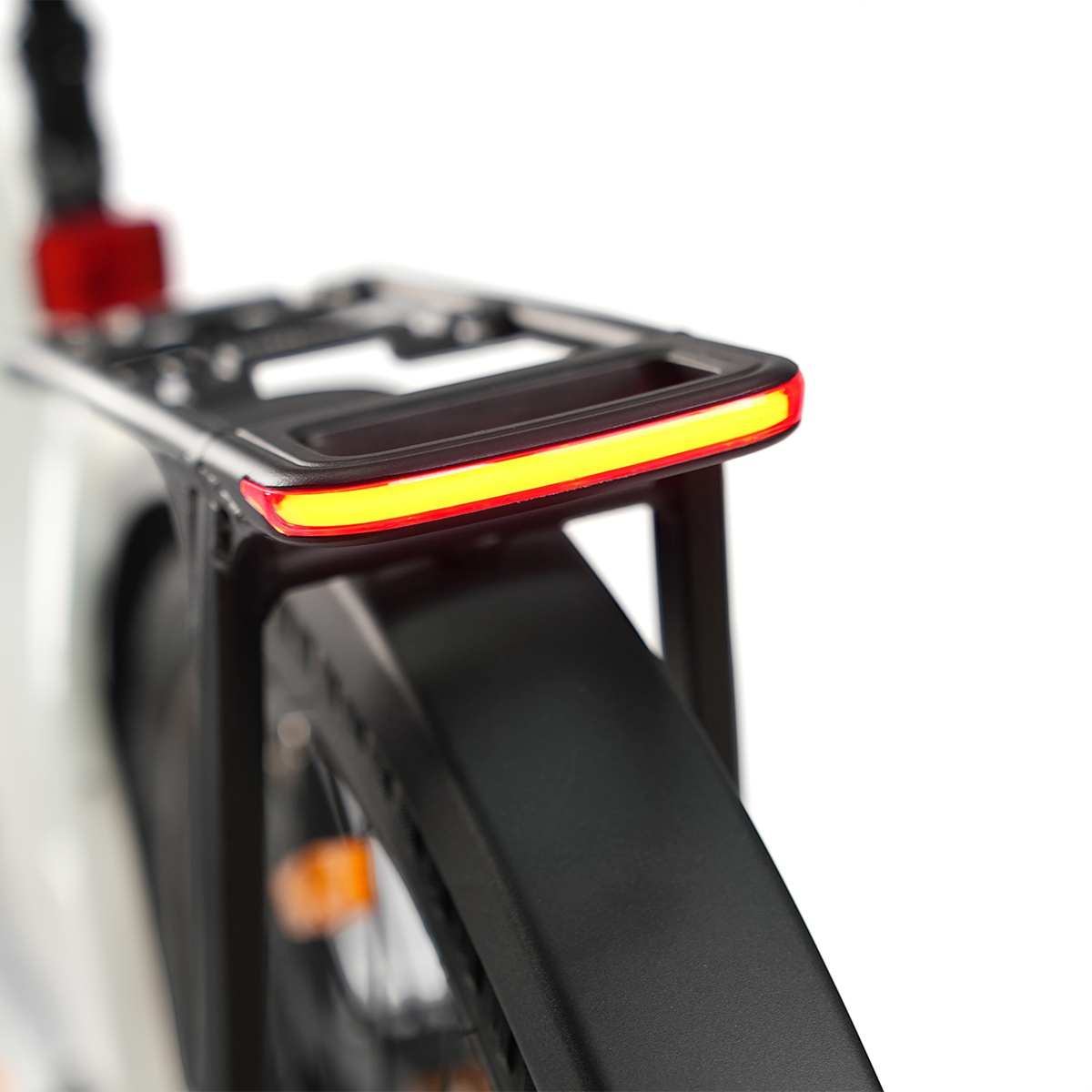 Tył roweru z czarnym bagażnikiem i jasnym, podświetlanym czerwonym i żółtym światłem. Widoczny jest czarny błotnik i część koła.