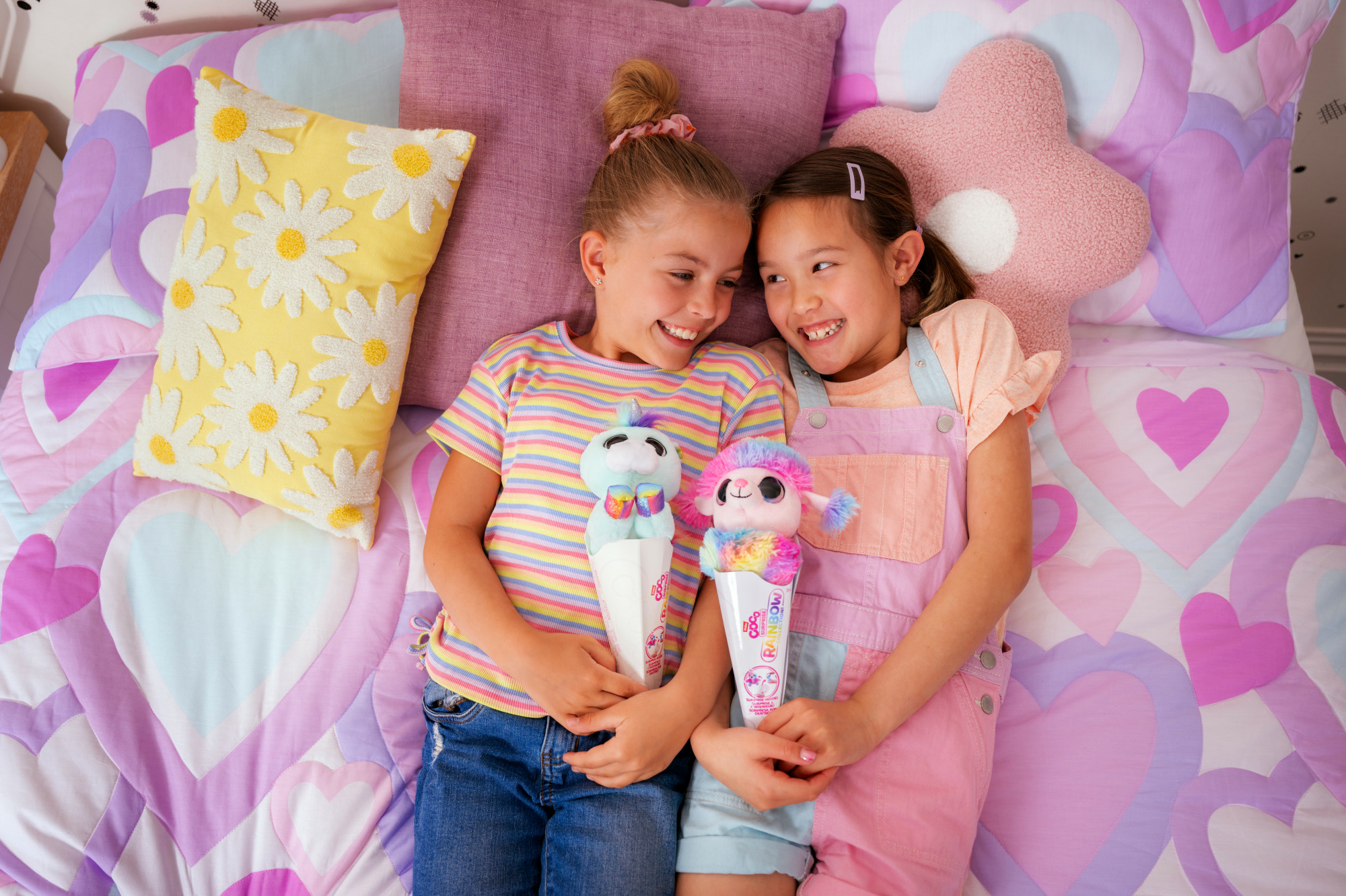 Due ragazze sorridenti sono sdraiate su un letto, tenendo in mano dei peluche. Il letto ha motivi a forma di cuore e cuscini. Una ragazza indossa una tuta.