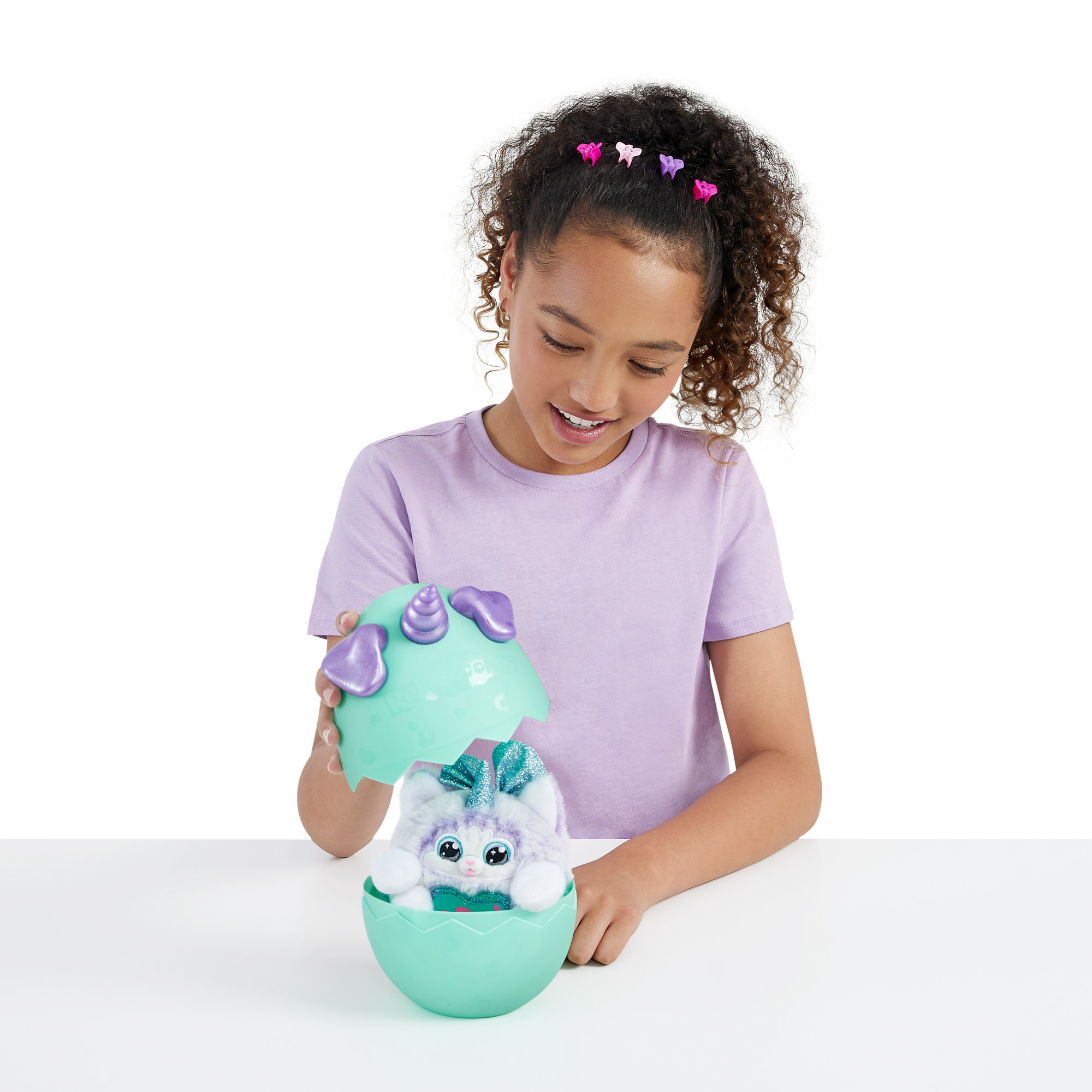 Dziewczyna otwiera turkusowe jajo, aby odsłonić pluszową zabawkę. Zabawka jest biała, fioletowa i ma błyszczącą kokardę.