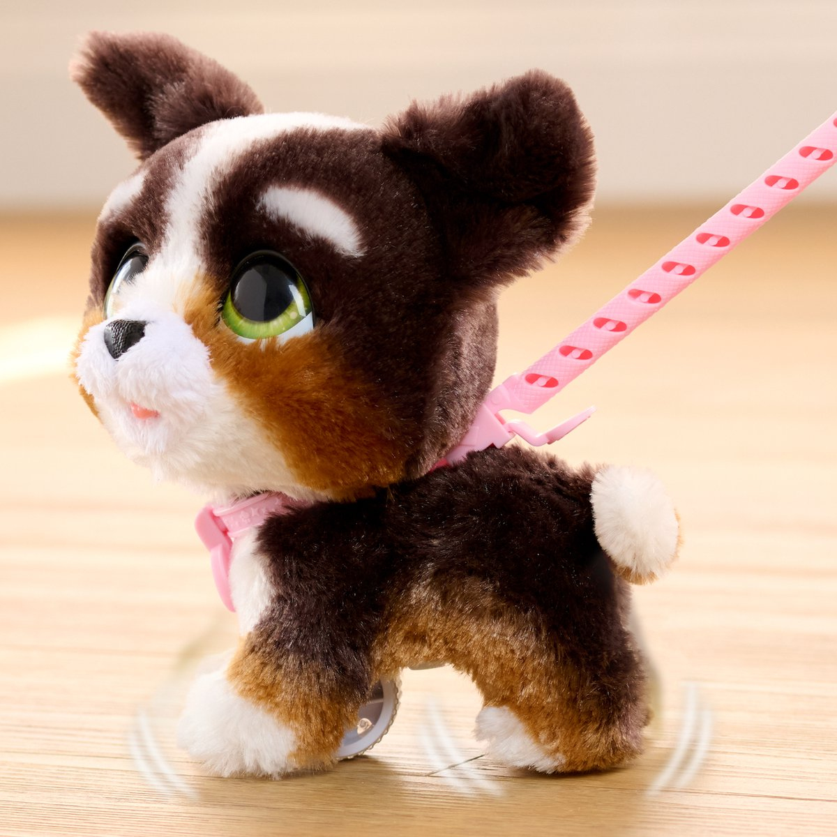 Un chien en peluche, marron, blanc et beige, avec de grands yeux verts et une laisse rose. Il est sur un sol en bois.