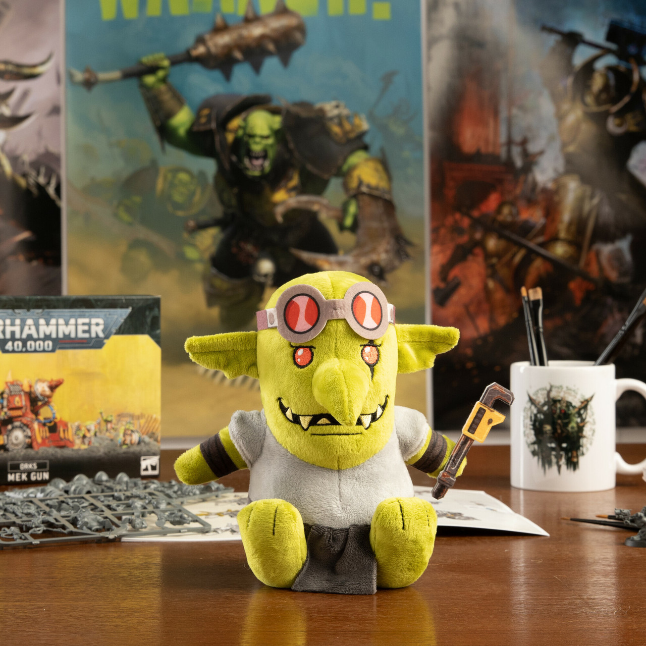 Un goblin di peluche verde con occhiali e una chiave inglese è seduto su un tavolo. Lo sfondo include l'arte di Warhammer, una tazza e parti di modellini.