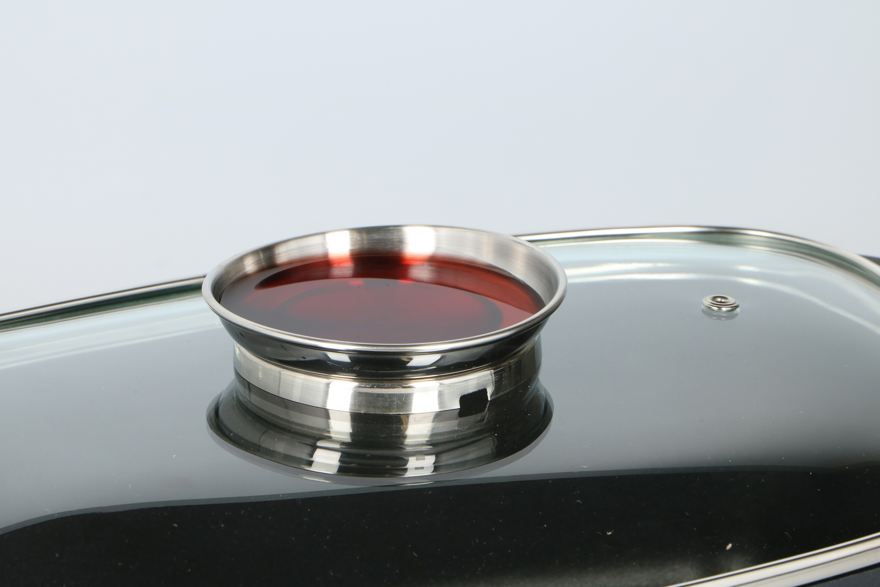 A close-up of a pot lid with a silver rim and a small, circular container with a red liquid inside.