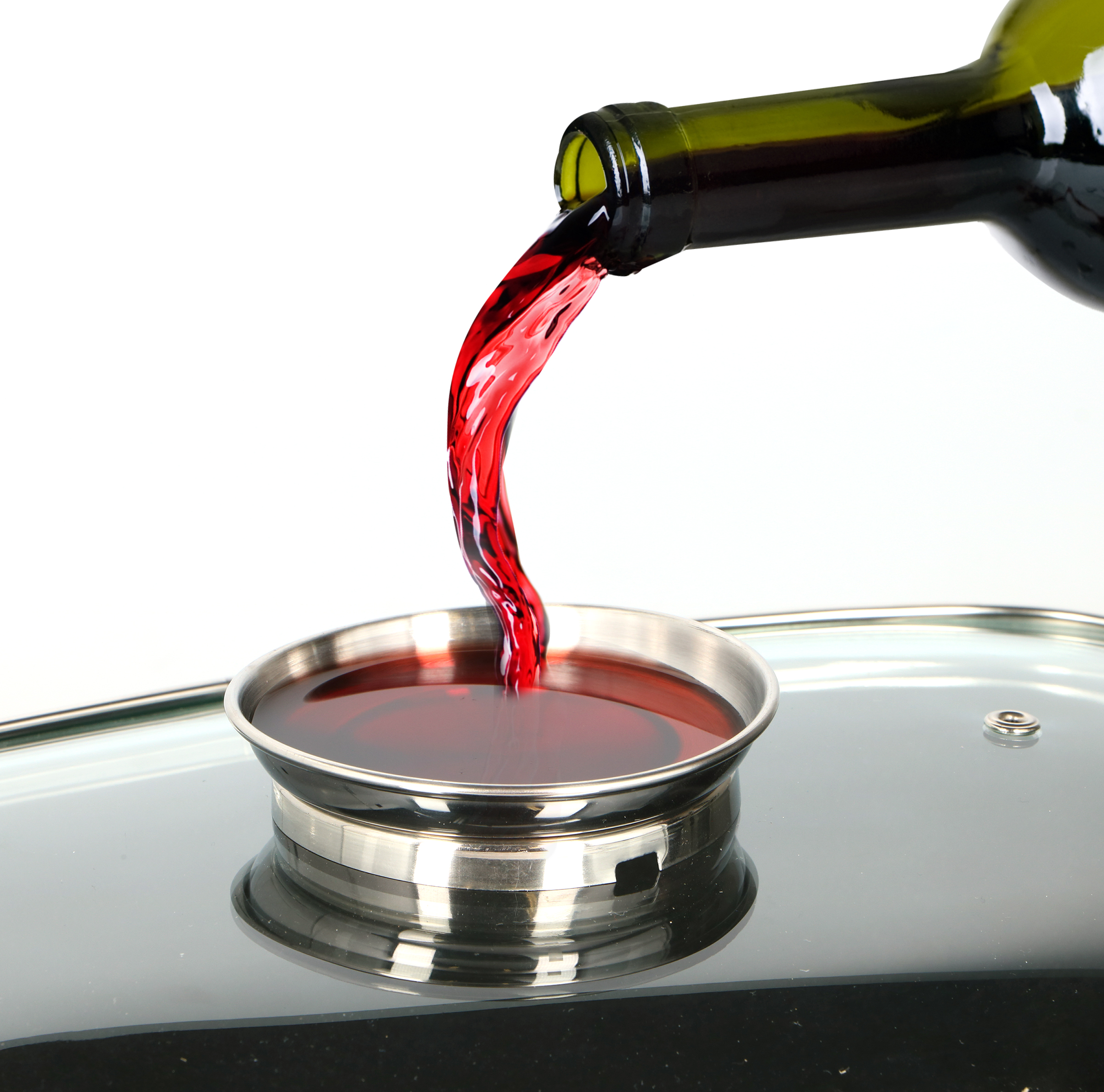 Red wine pouring from a green bottle into a small silver dish. The dish sits on a glass surface.