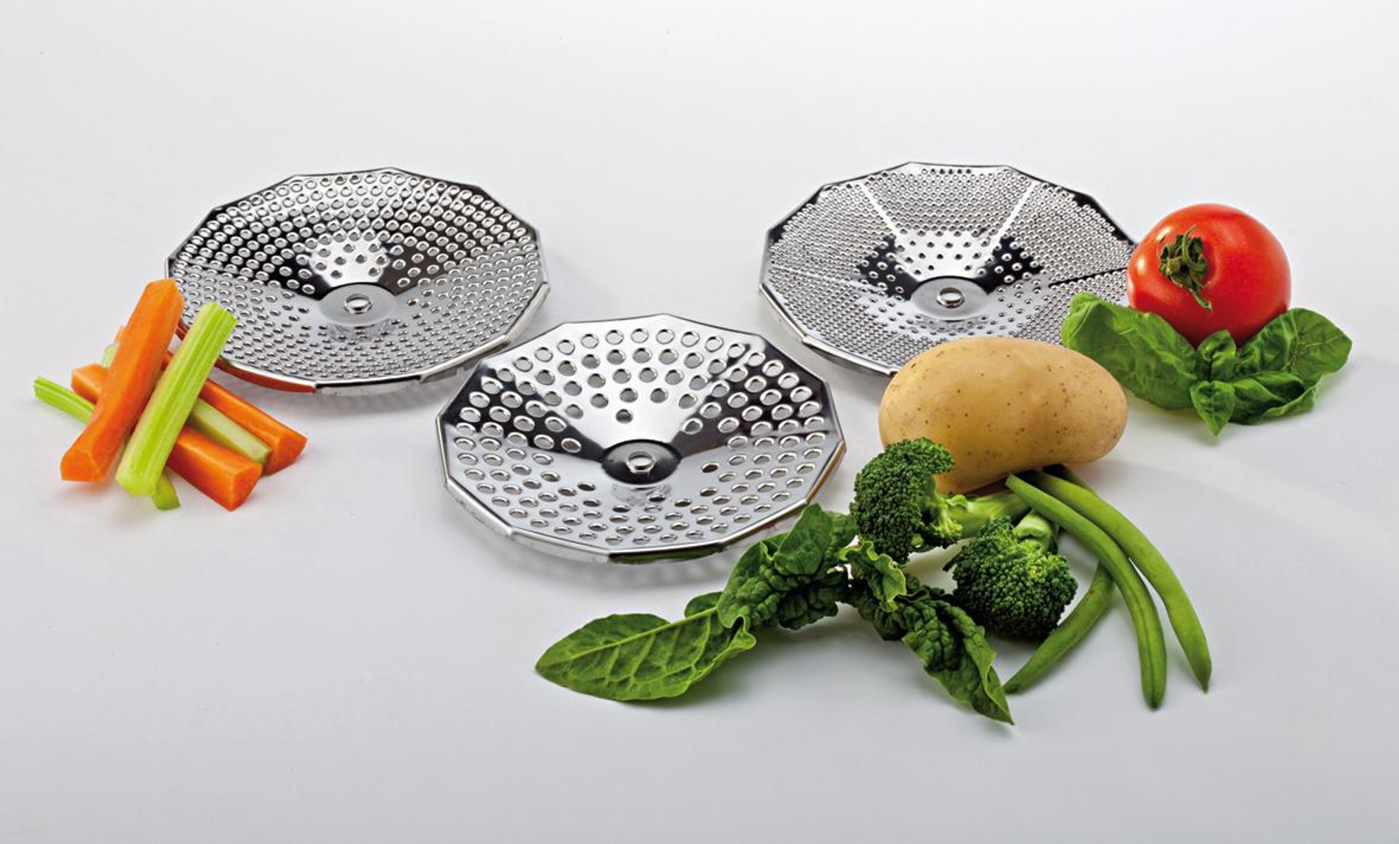 Drei Metall-Dampfgarerkörbe umgeben Gemüse wie Karotten, Tomaten, Kartoffeln, Brokkoli und Bohnen auf weißer Oberfläche.