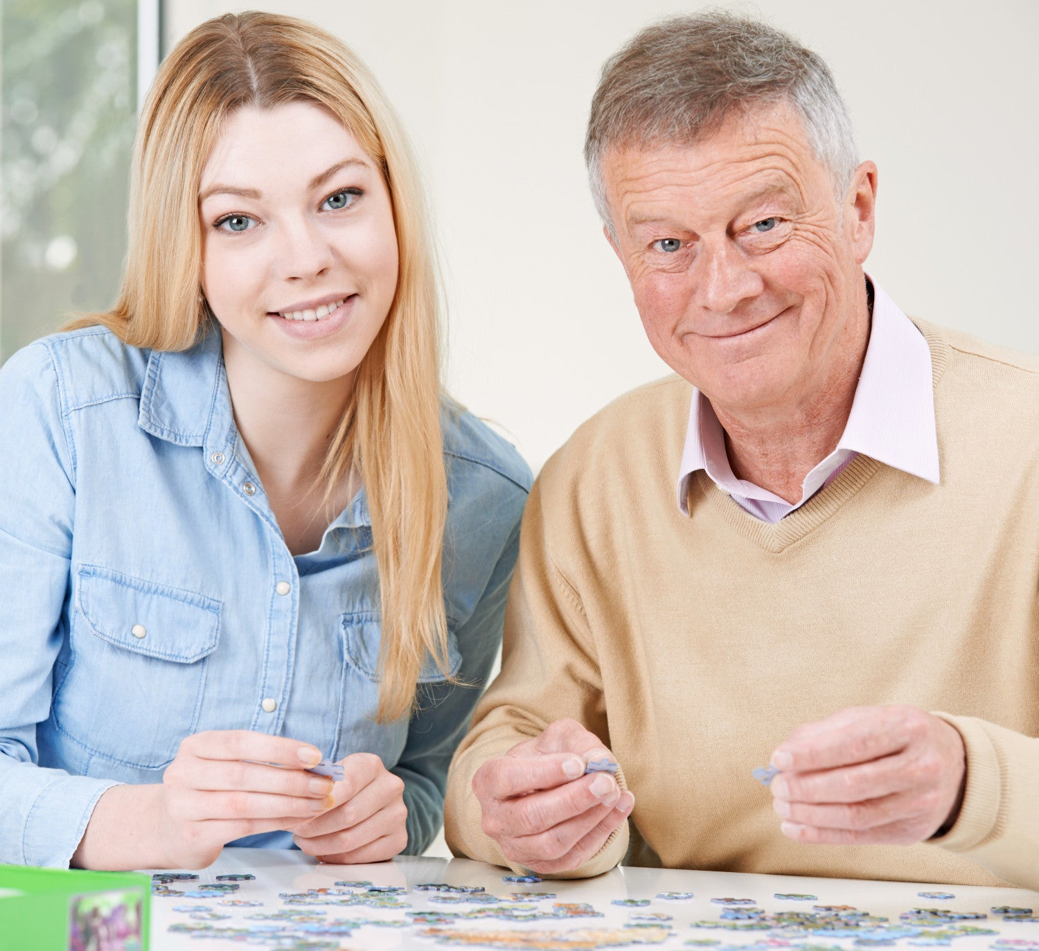 Eine junge Frau und ein älterer Mann sitzen an einem Tisch und setzen ein Puzzle zusammen. Beide lächeln und halten Puzzleteile.