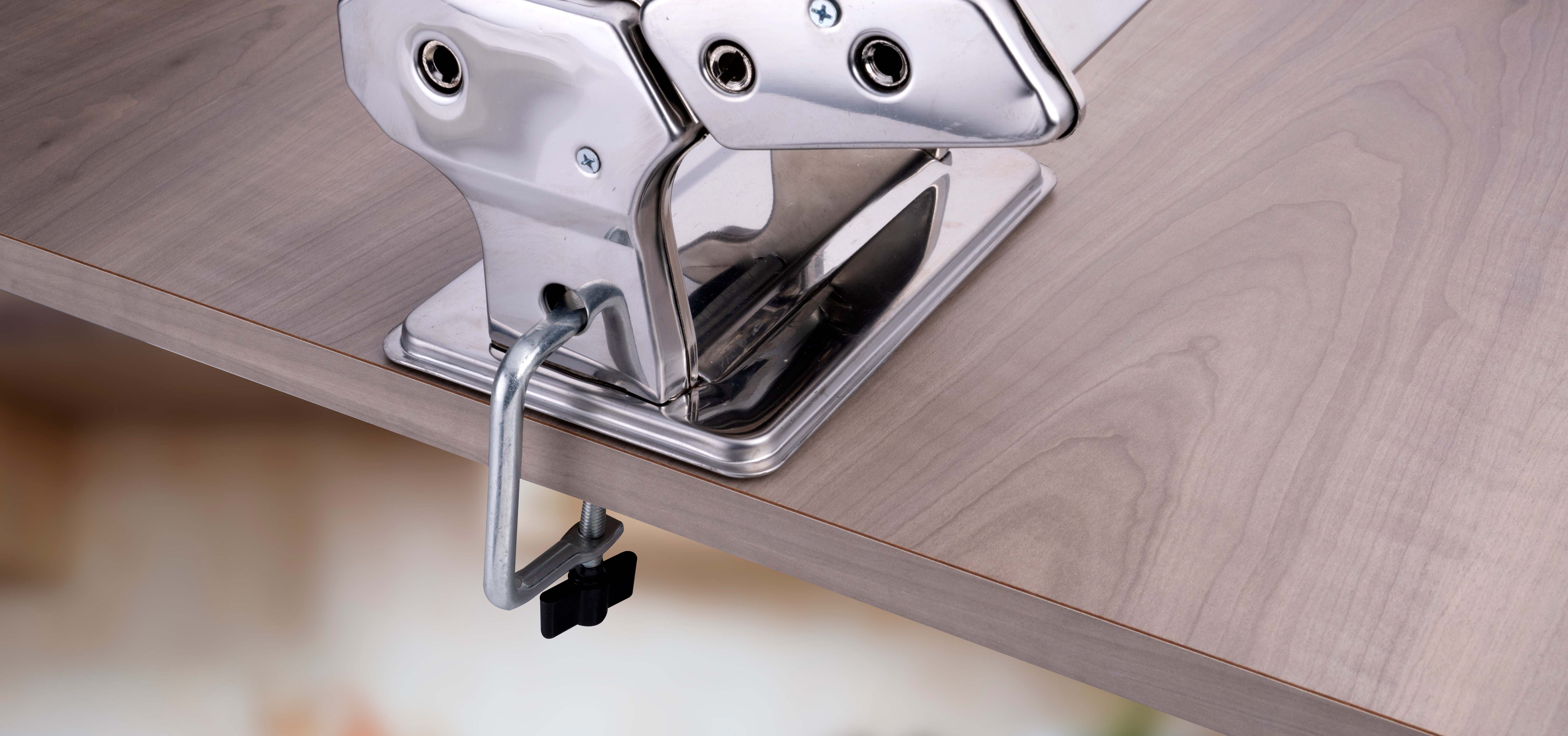 A close-up of a pasta machine clamped to a wooden surface. The machine is silver, and the wood is light brown.