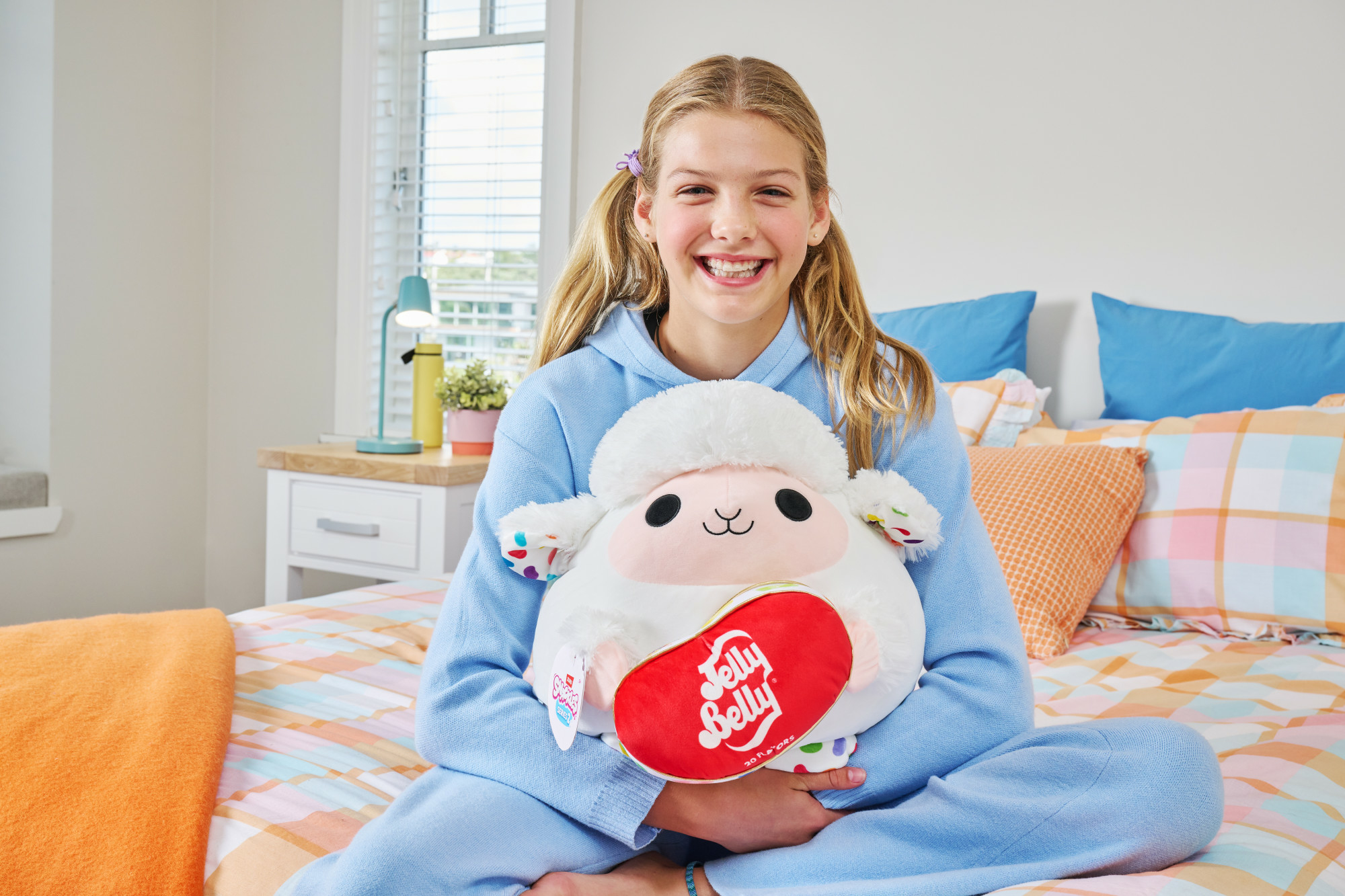 Ein lächelndes Mädchen hält ein Schaf-Plüschtier mit Jelly Belly Logo. Sie sitzt auf einem Bett mit bunter Bettwäsche. Im Hintergrund ein Fenster.