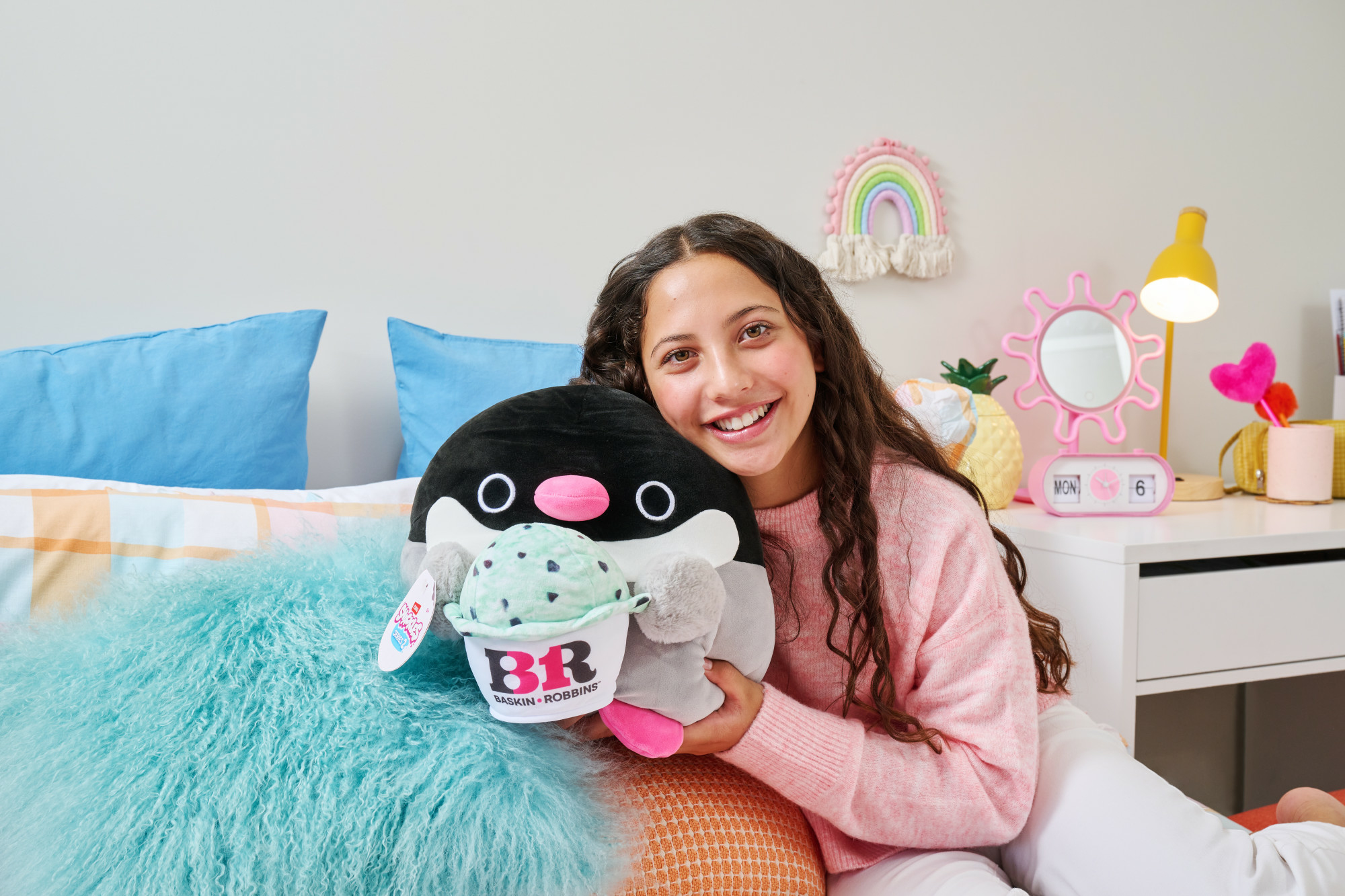Una ragazza sorridente tiene in mano un peluche a forma di pinguino con una coppetta gelato. È seduta su un letto con dei cuscini. C'è una decorazione arcobaleno sul muro.
