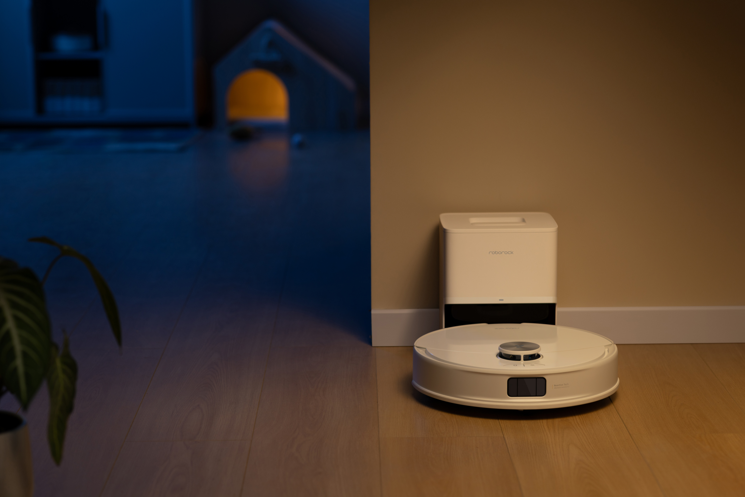 Een witte robotstofzuiger en zijn oplaadstation op een houten vloer. Een plant en een speelgoedhuis op de achtergrond.