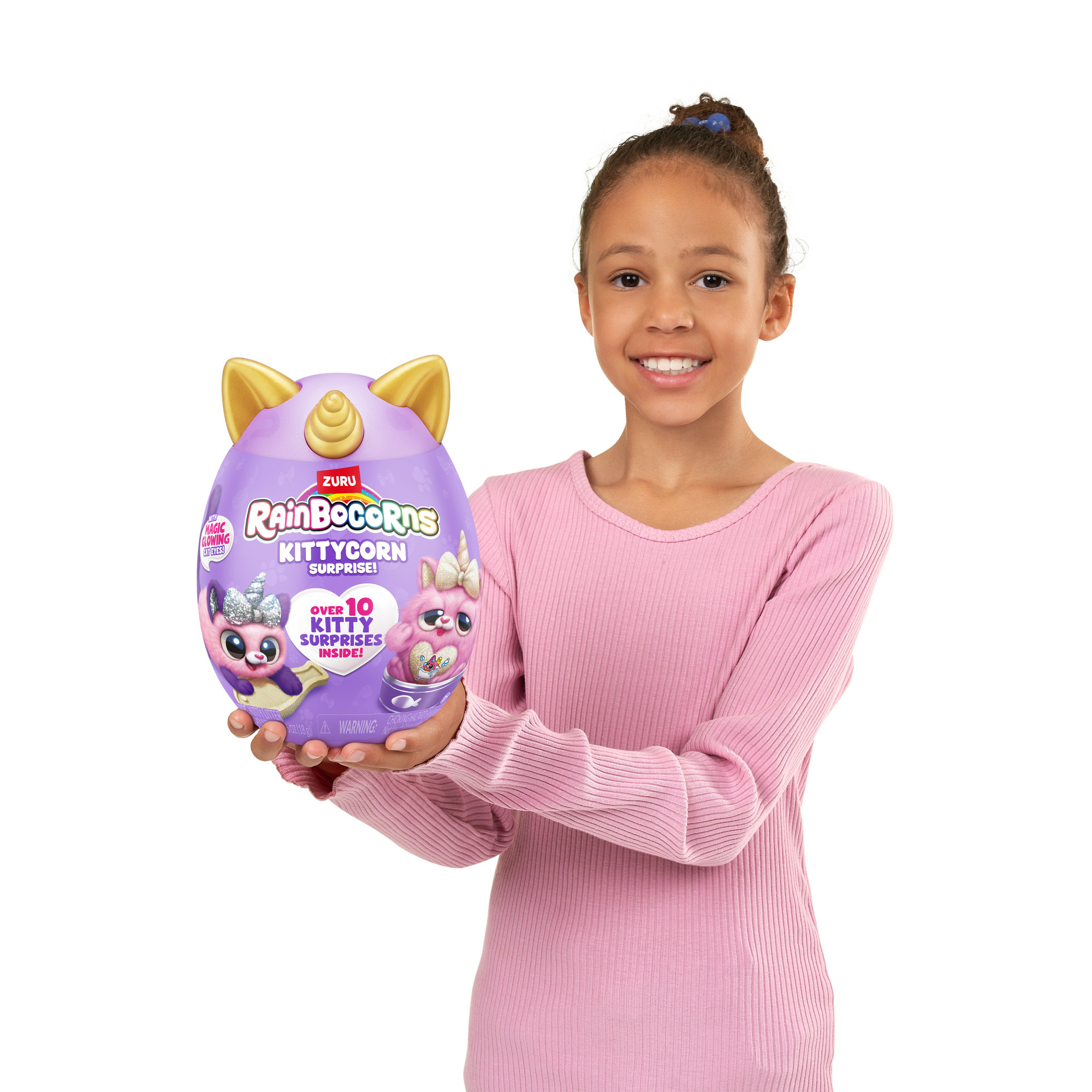 Ein Mädchen hält ein lila, eiförmiges Spielzeug mit Einhorn-Details. Das Spielzeug trägt die Aufschrift Rainbocorns Kittycorn Surprise.