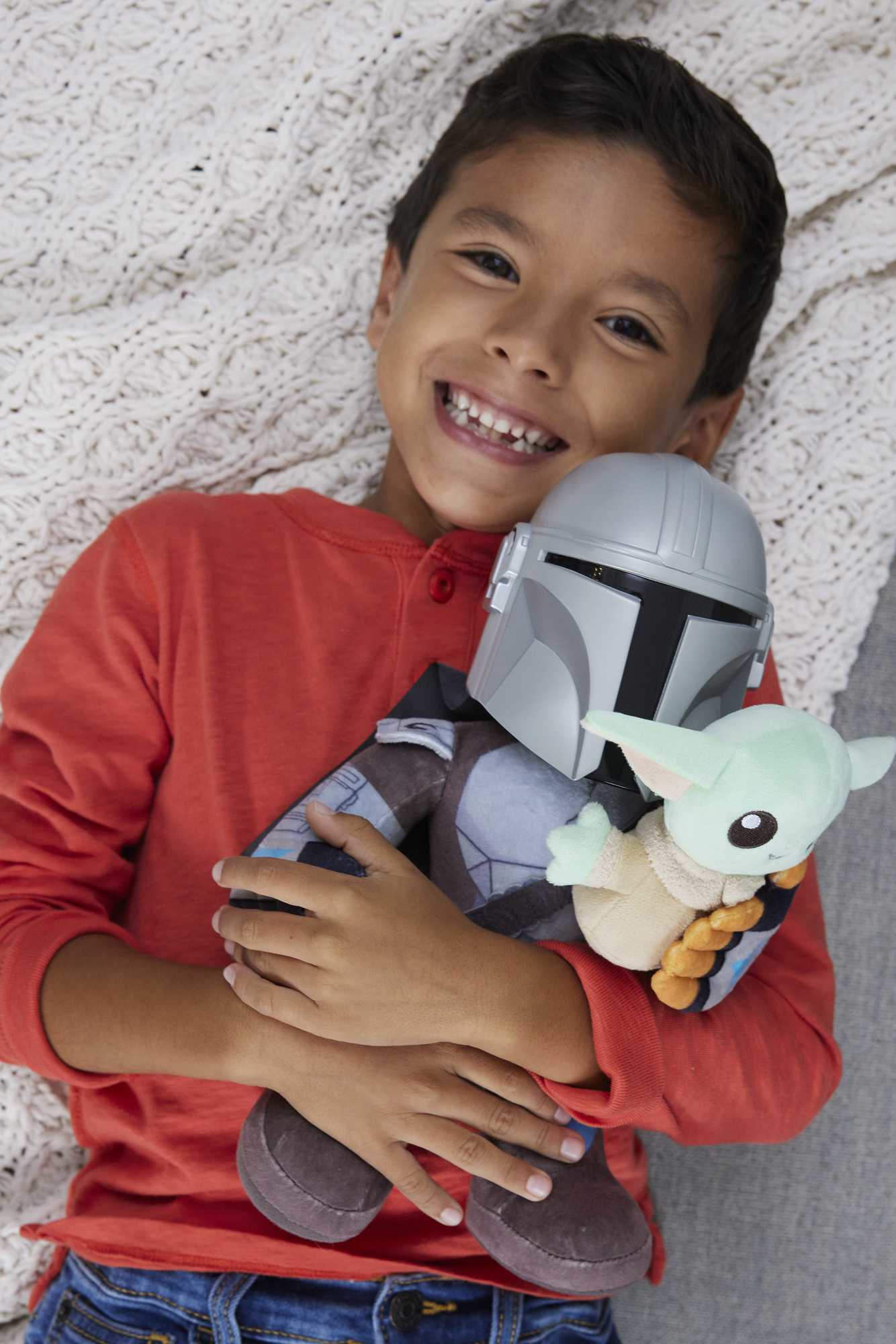 Uśmiechnięty chłopiec w czerwonej koszuli trzyma pluszaki Mandalorian i Grogu. W tle biały koc.