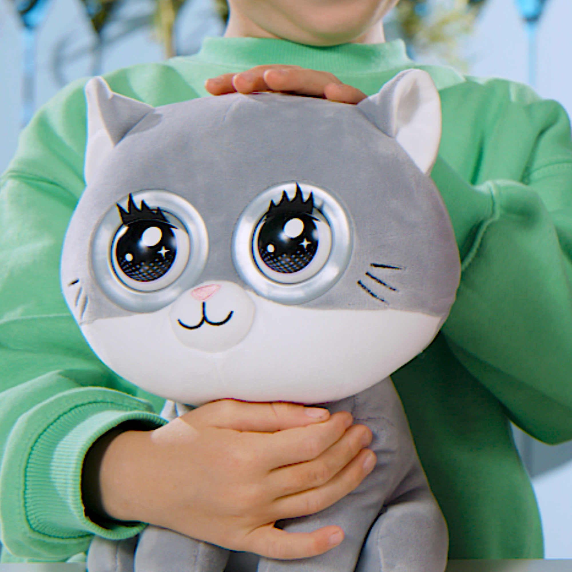 Un bambino tiene in mano un peluche di gatto grigio con grandi occhi. Il bambino indossa un maglione verde.