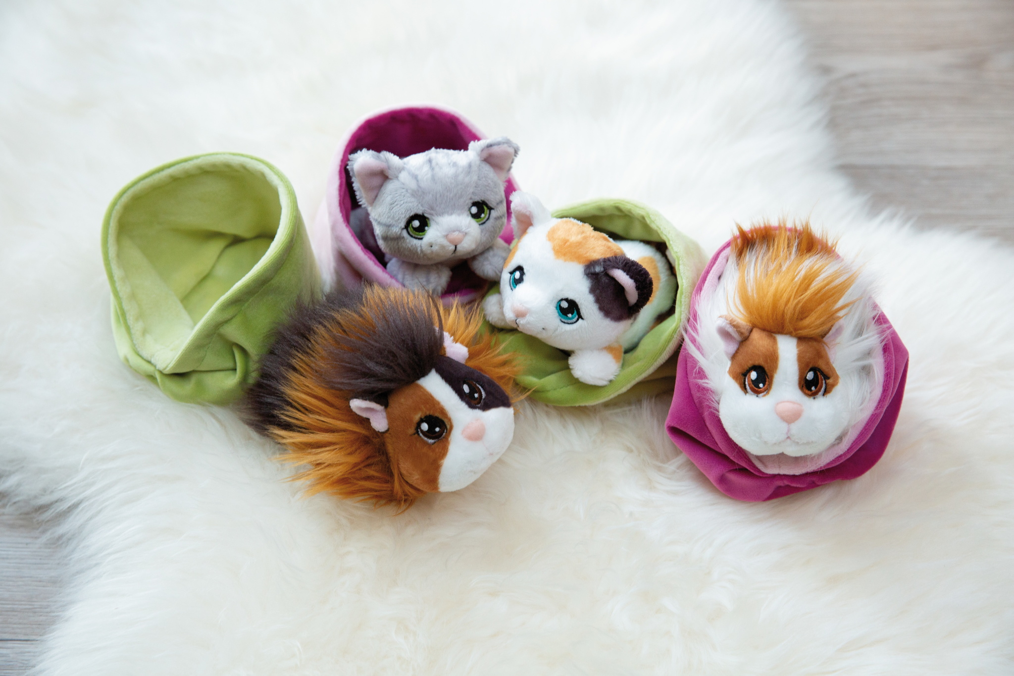 Vier Plüschtiere, eine graue Katze, eine Schildpattkatze und zwei Meerschweinchen, in farbenfrohen, stoffgefütterten Behältern auf weißer, flauschiger Oberfläche.