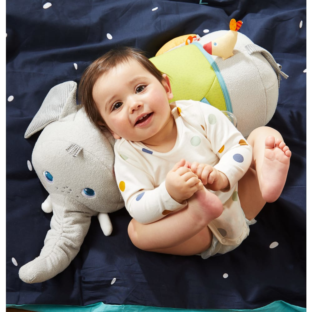 Ein Baby in einem gepunkteten Strampler liegt auf einer marineblauen Decke mit Spielzeug. Ein Elefant und ein rundes Spielzeug liegen auf der Decke.