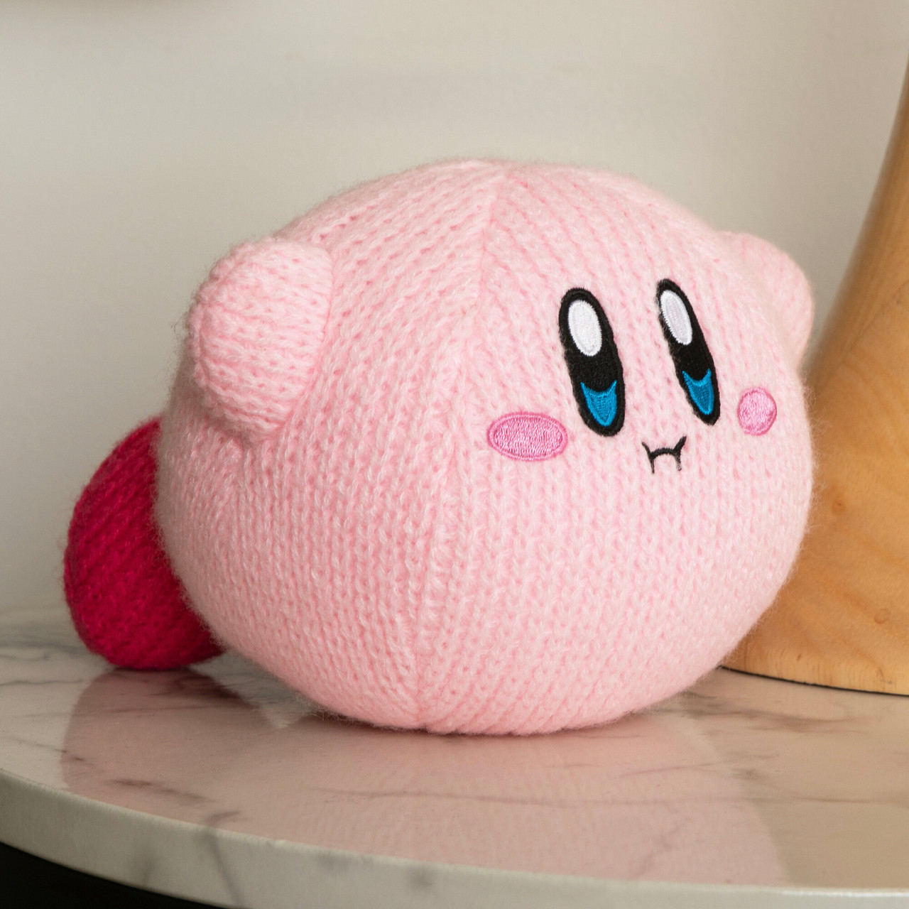 Una bambola Kirby rosa lavorata a maglia con occhi neri e riflessi blu, un piccolo sorriso e piedi rossi è su una superficie bianca.