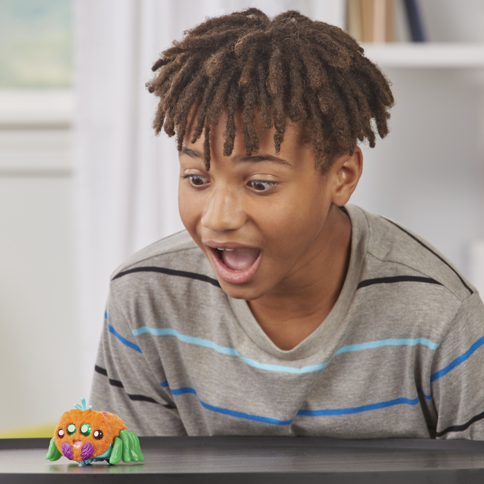 Ein Junge mit Dreadlocks blickt überrascht auf eine Spielzeugspinne. Das Spielzeug ist orange, grün und lila. Der Junge trägt ein graues Hemd.
