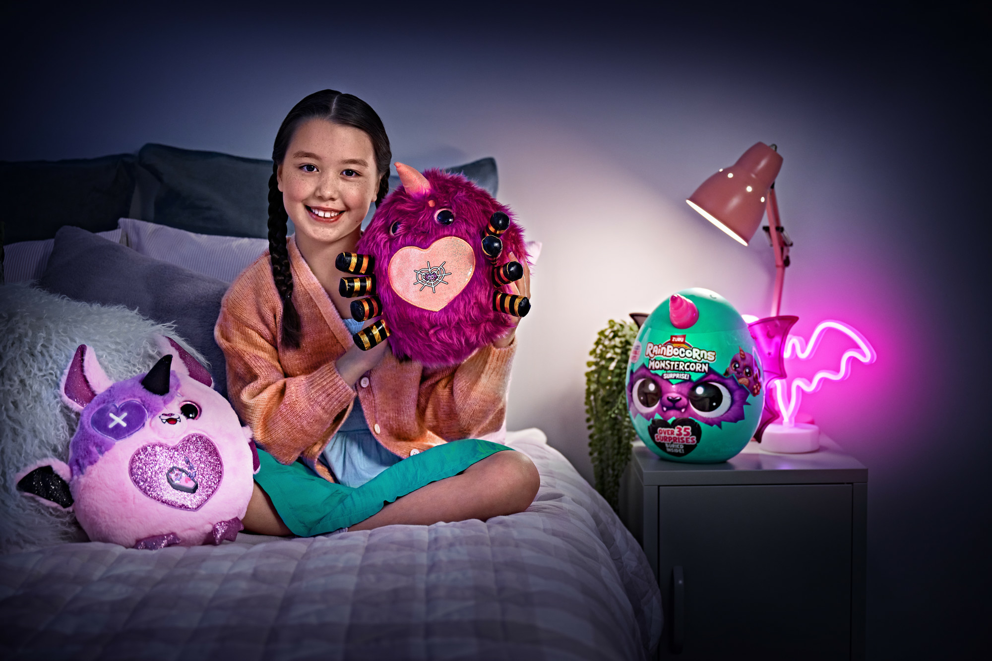 Una ragazza sorride, tenendo in mano un peluche rosa. Accanto a lei ci sono altri due giocattoli. Un pipistrello al neon e una lampada sono su un comodino.