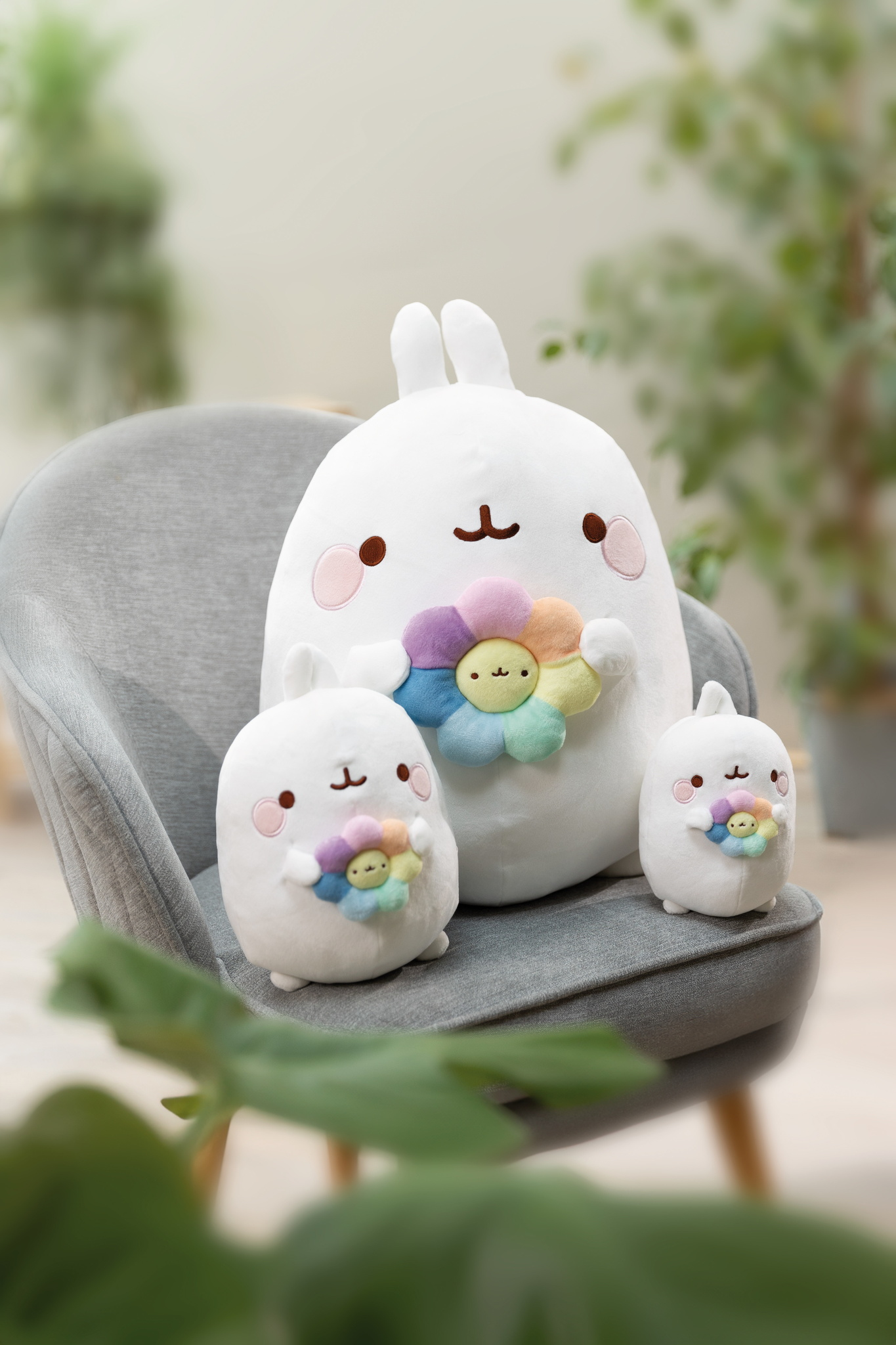 Tre peluche di coniglietti bianchi su una sedia grigia, che tengono peluche di fiori colorati. Piante verdi sullo sfondo.