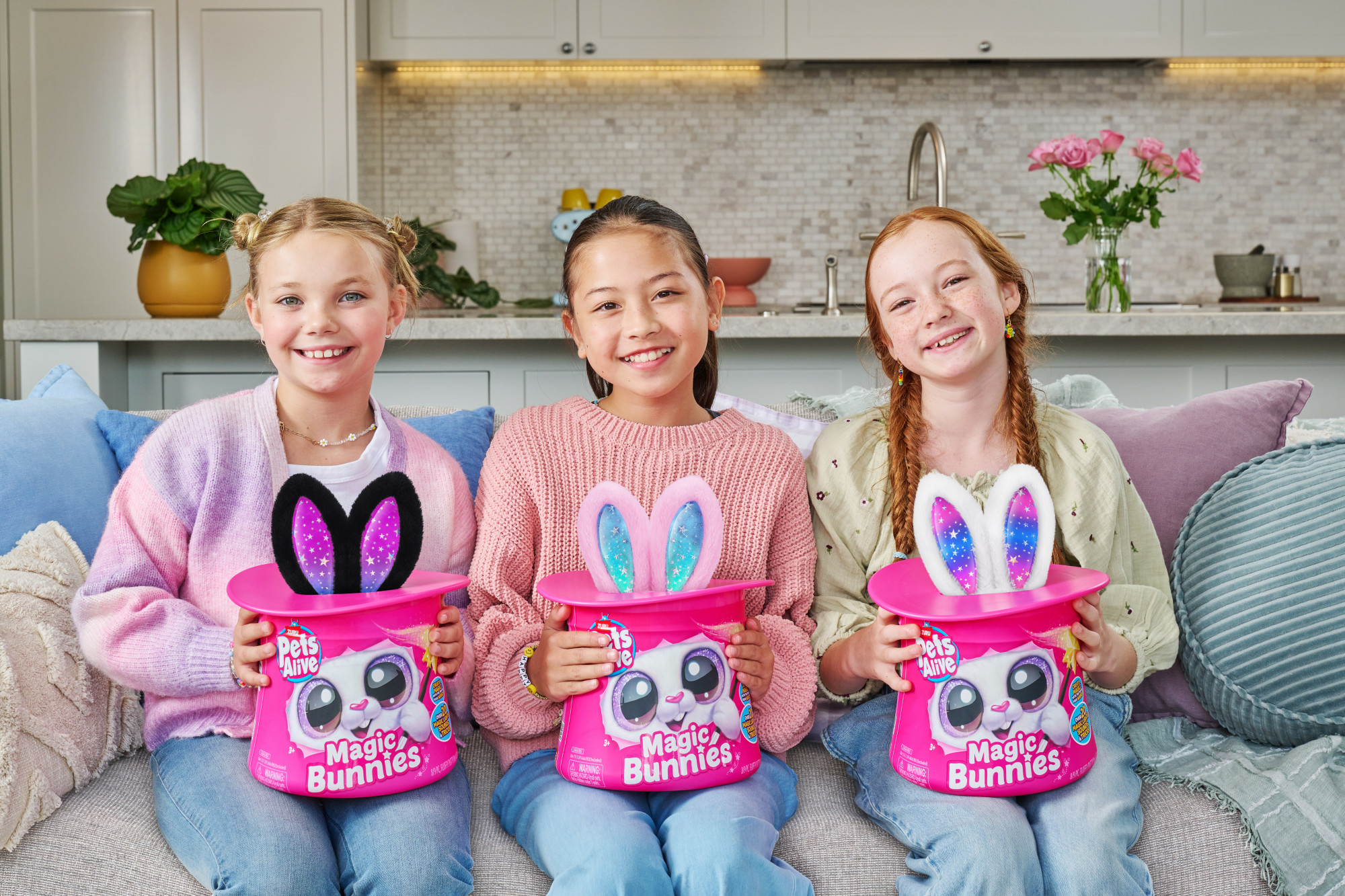 Drei Mädchen halten rosa 'Magic Bunnies'-Boxen mit Hasenohren. Sie lächeln und sitzen auf einer Couch mit Kissen.