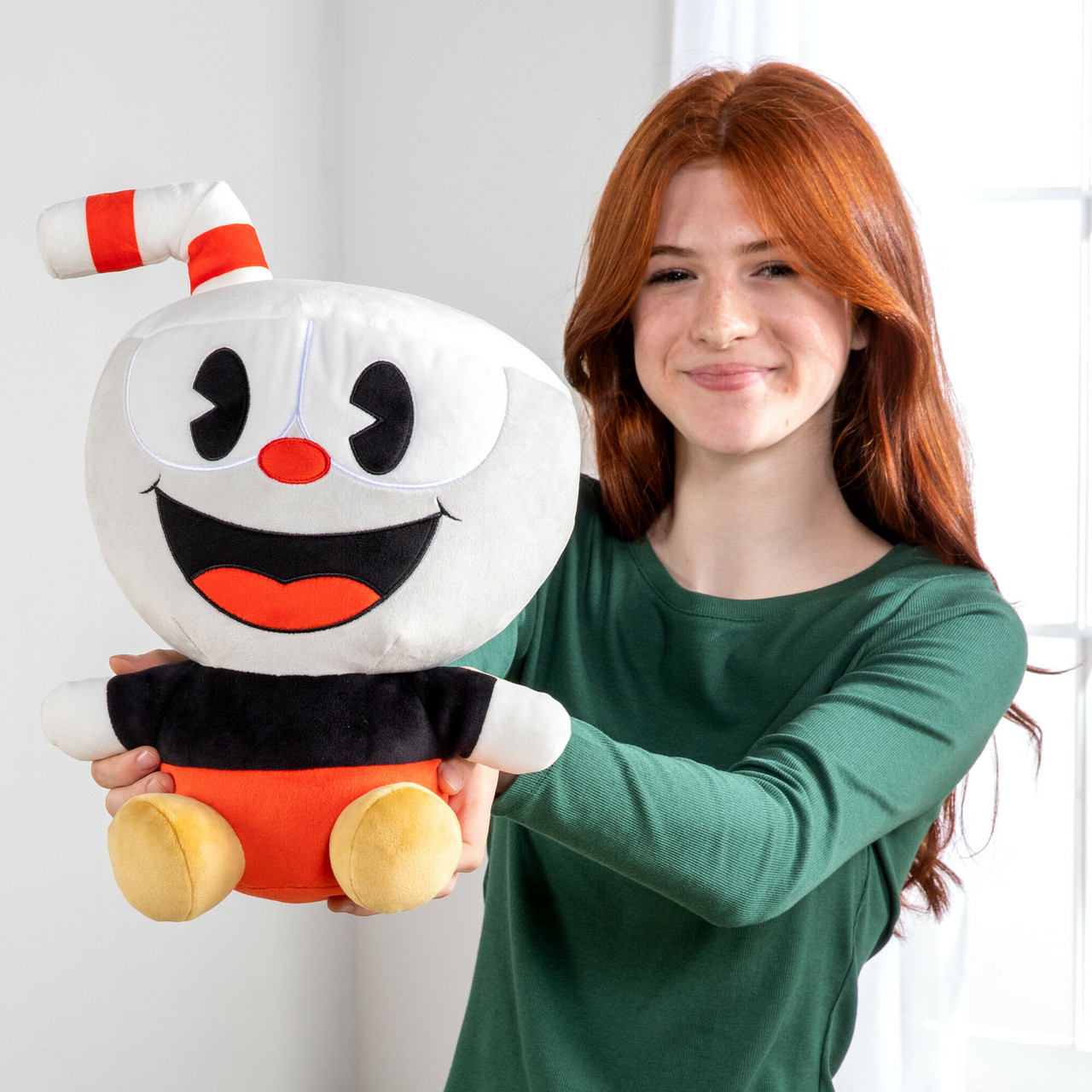 Una ragazza dai capelli rossi sorridente tiene in mano un peluche di Cuphead. Il giocattolo è bianco con occhi neri e naso rosso. La ragazza indossa una camicia verde.