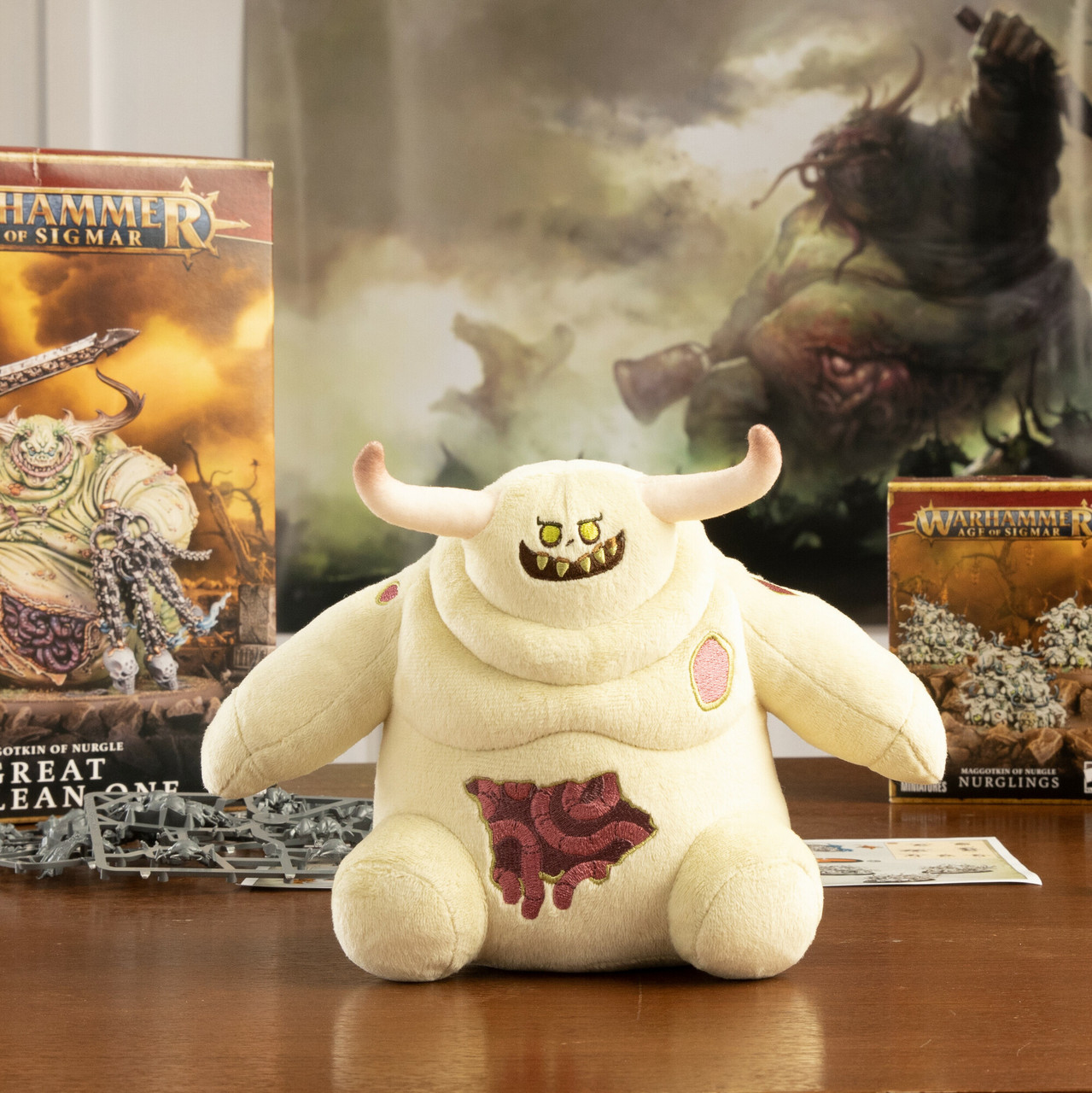 Une peluche d'une créature avec des cornes et un visage souriant est sur une table. Des boîtes Warhammer sont en arrière-plan.